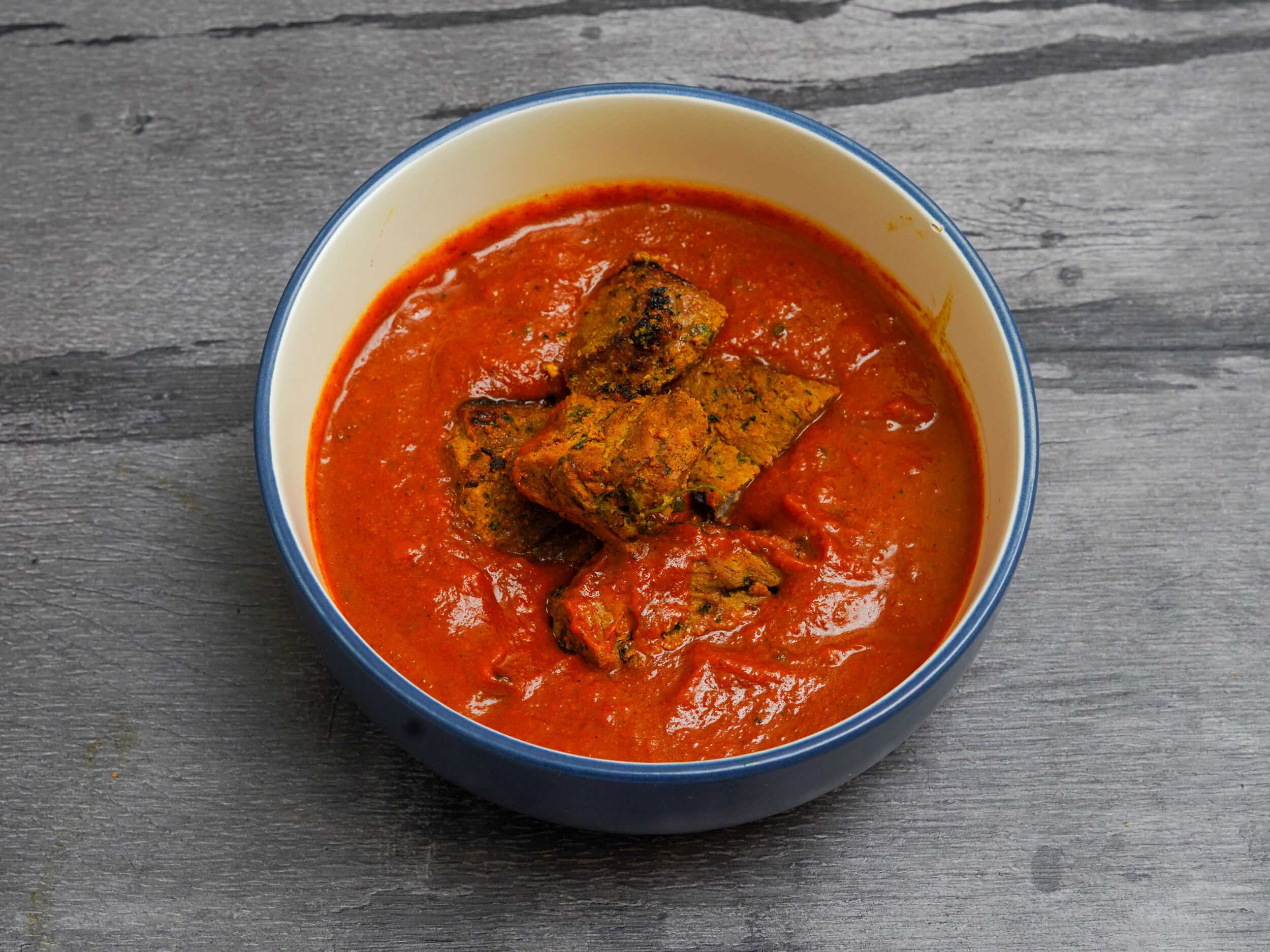 Four pieces of marinated chicken or paneer in a vivid red-orange curry sauce in a blue-rimmed bowl on a grey wooden surface