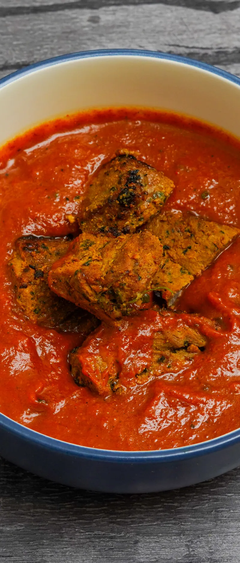 Four pieces of marinated chicken or paneer in a vivid red-orange curry sauce in a blue-rimmed bowl on a grey wooden surface