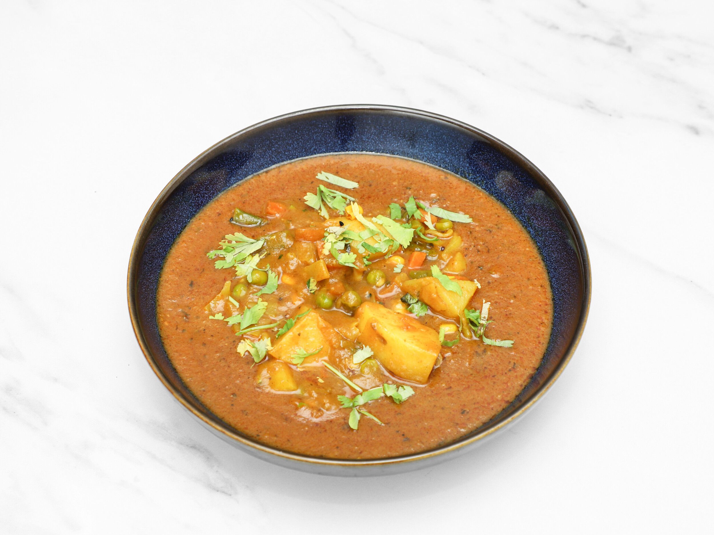 A bowl of vegetable curry with potatoes, green peas, carrots, and cilantro on a white marble surface.