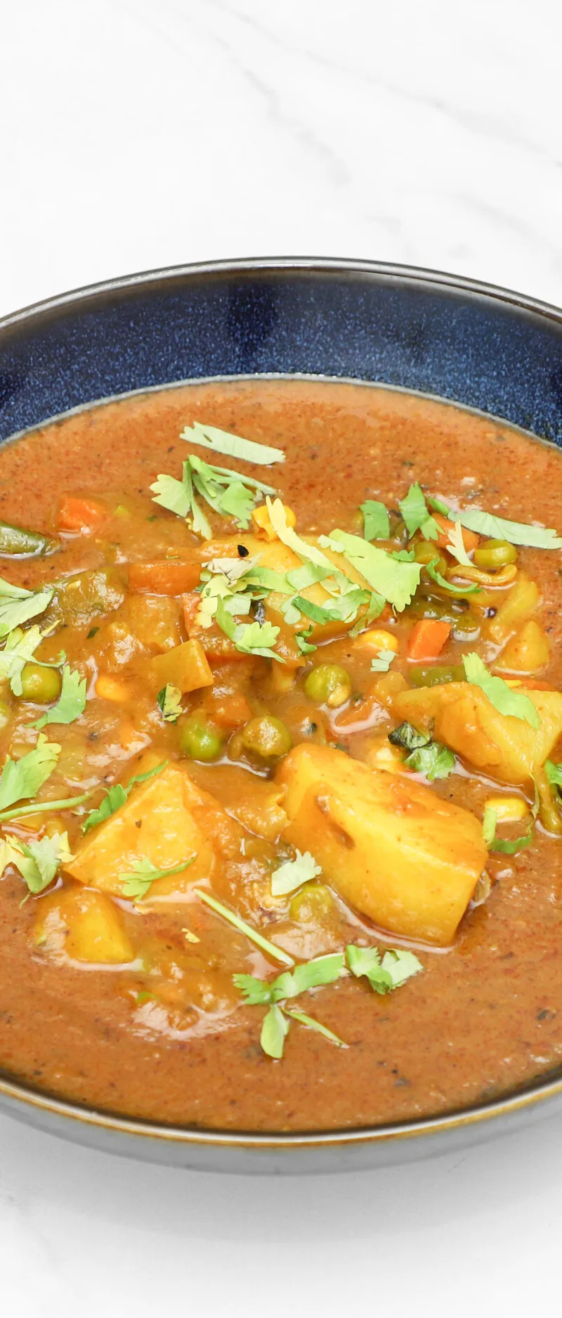 A bowl of vegetable curry with potatoes, green peas, carrots, and cilantro on a white marble surface.