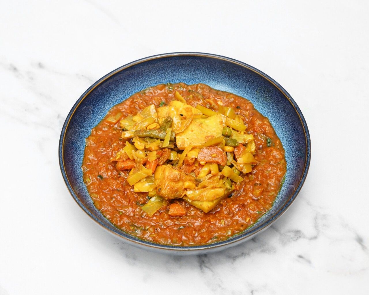 Top-down view of a colorful vegetable and chicken curry in a deep blue bowl on a white surface.