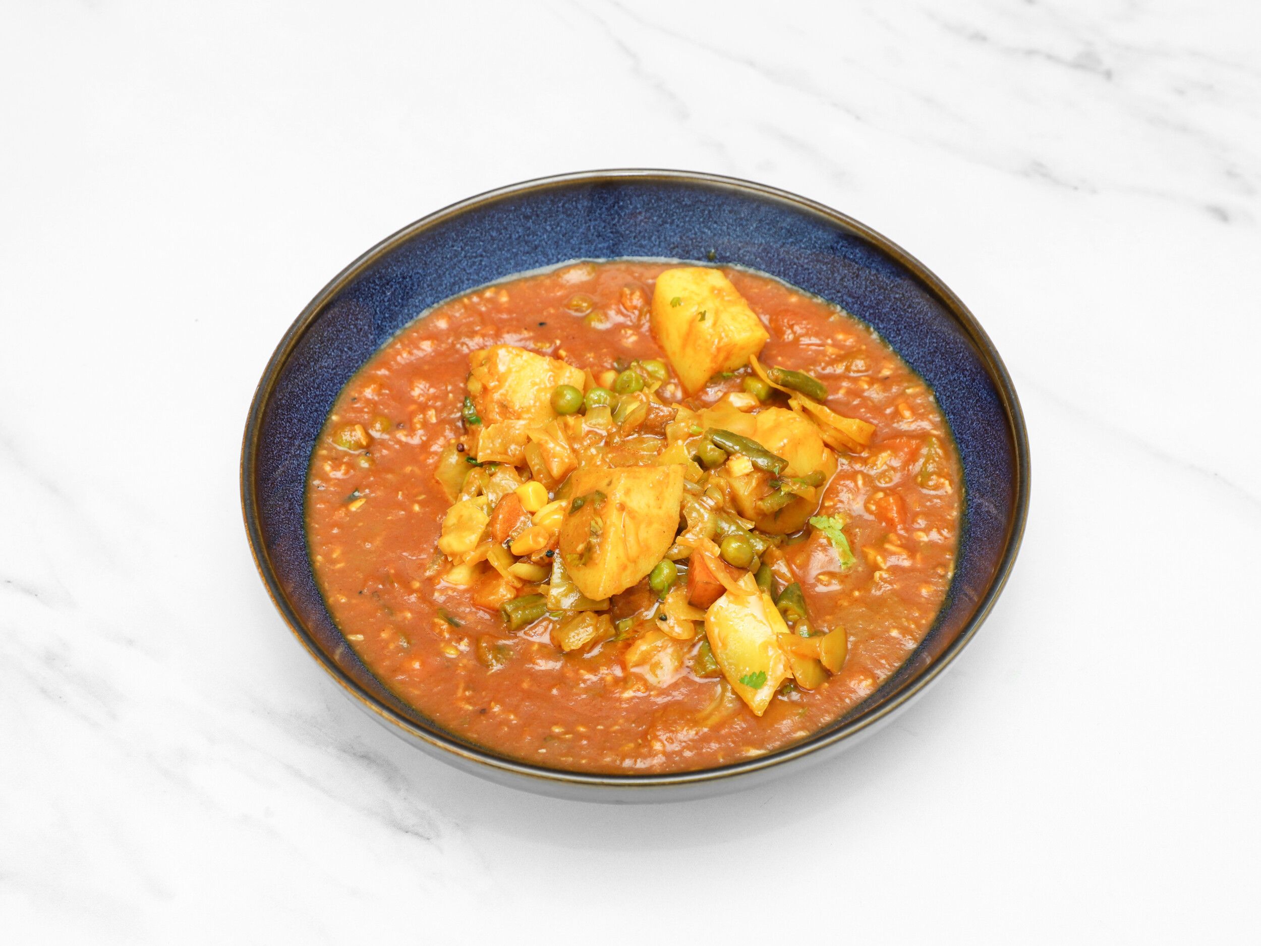 A close-up of a blue bowl filled with a rich orange-red vegetable curry on a white marble surface.