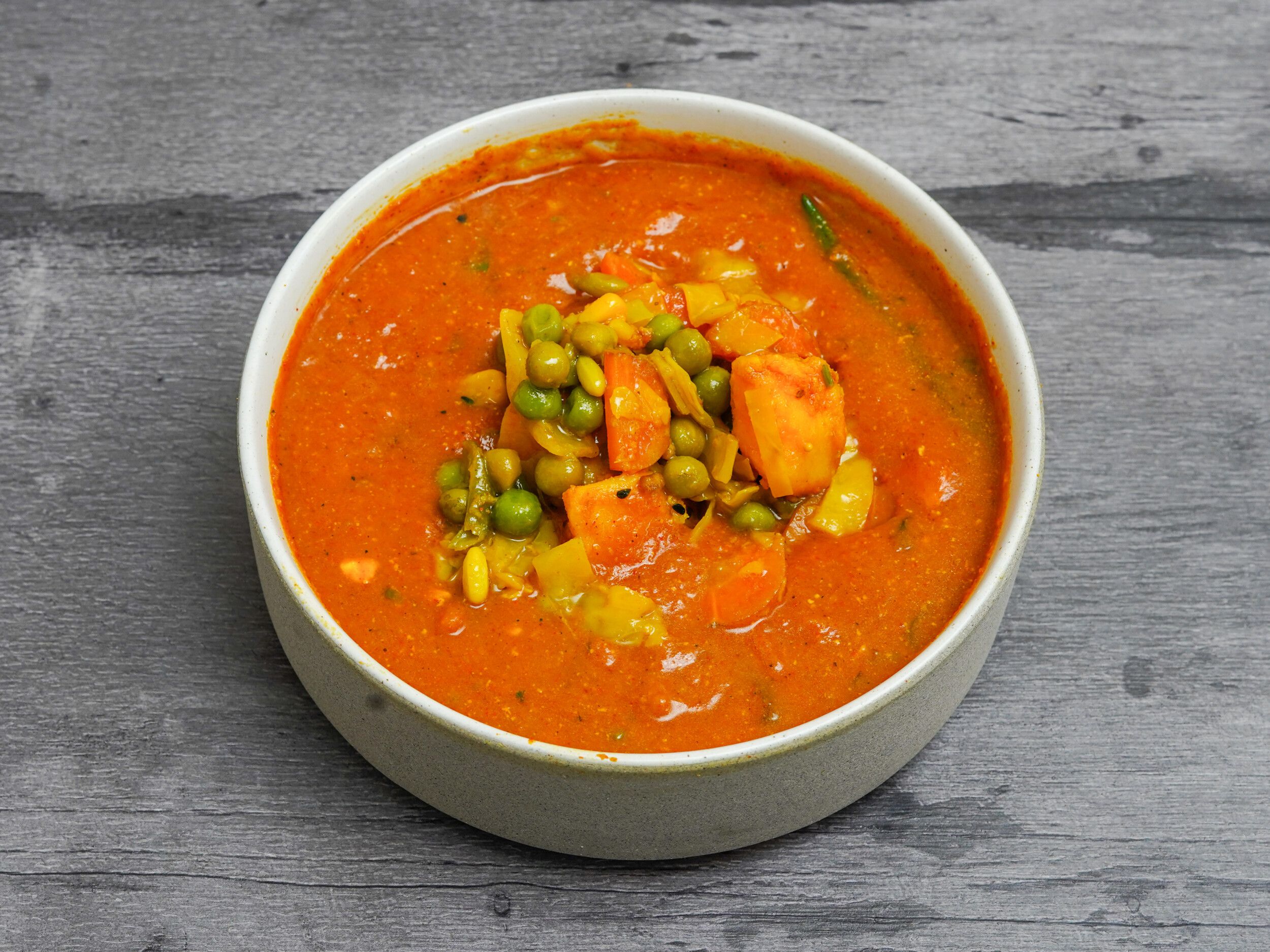 A white bowl filled with an orange vegetable curry, topped with green peas and diced carrots.