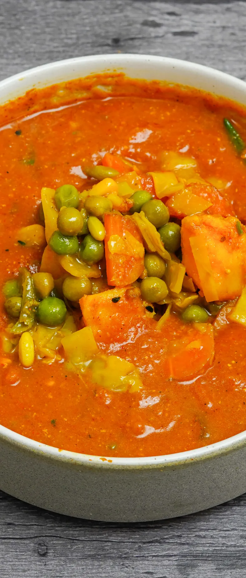 A white bowl filled with an orange vegetable curry, topped with green peas and diced carrots.