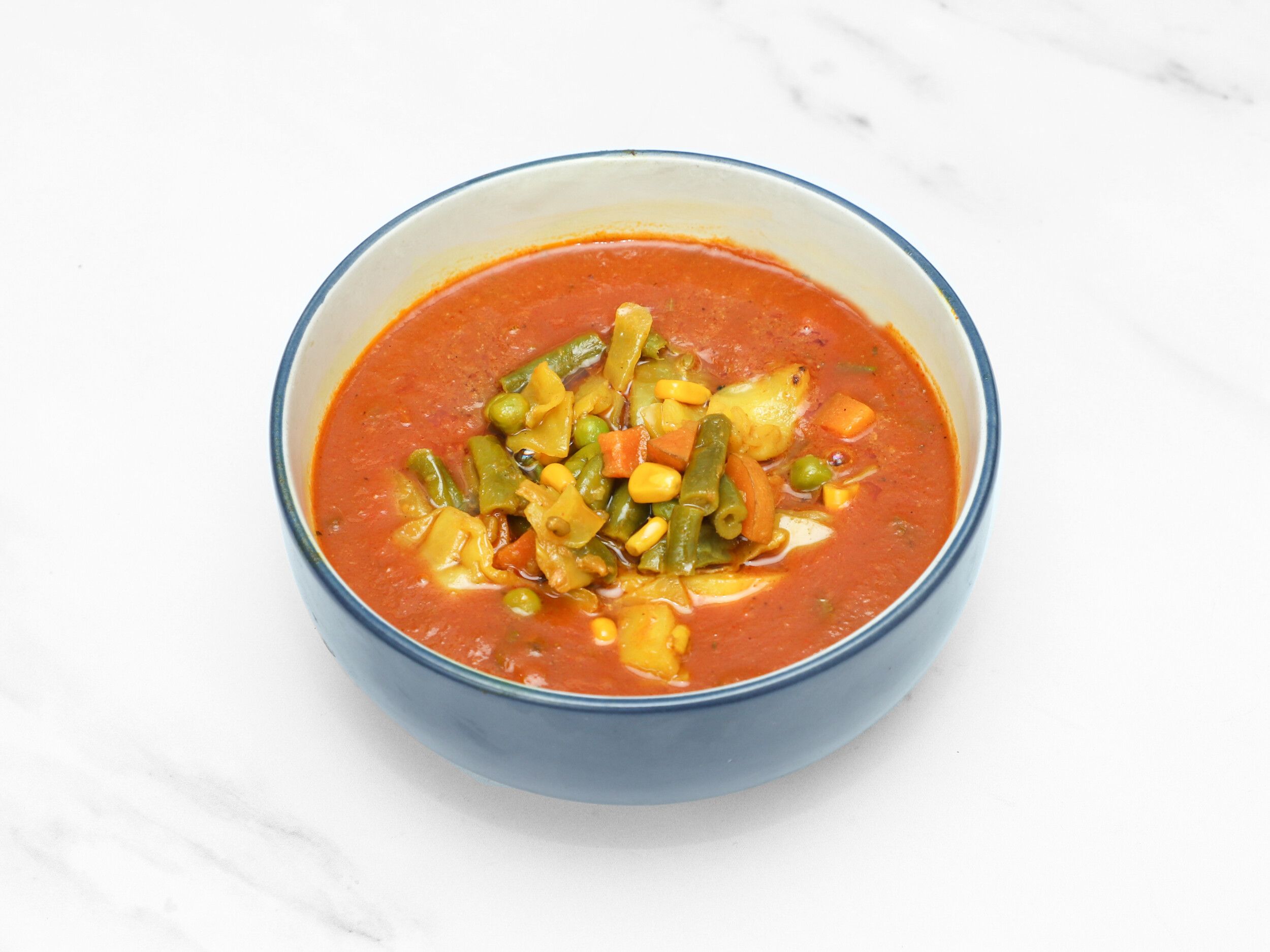 A close-up of mixed vegetable curry in a blue ceramic bowl on a white marble surface.