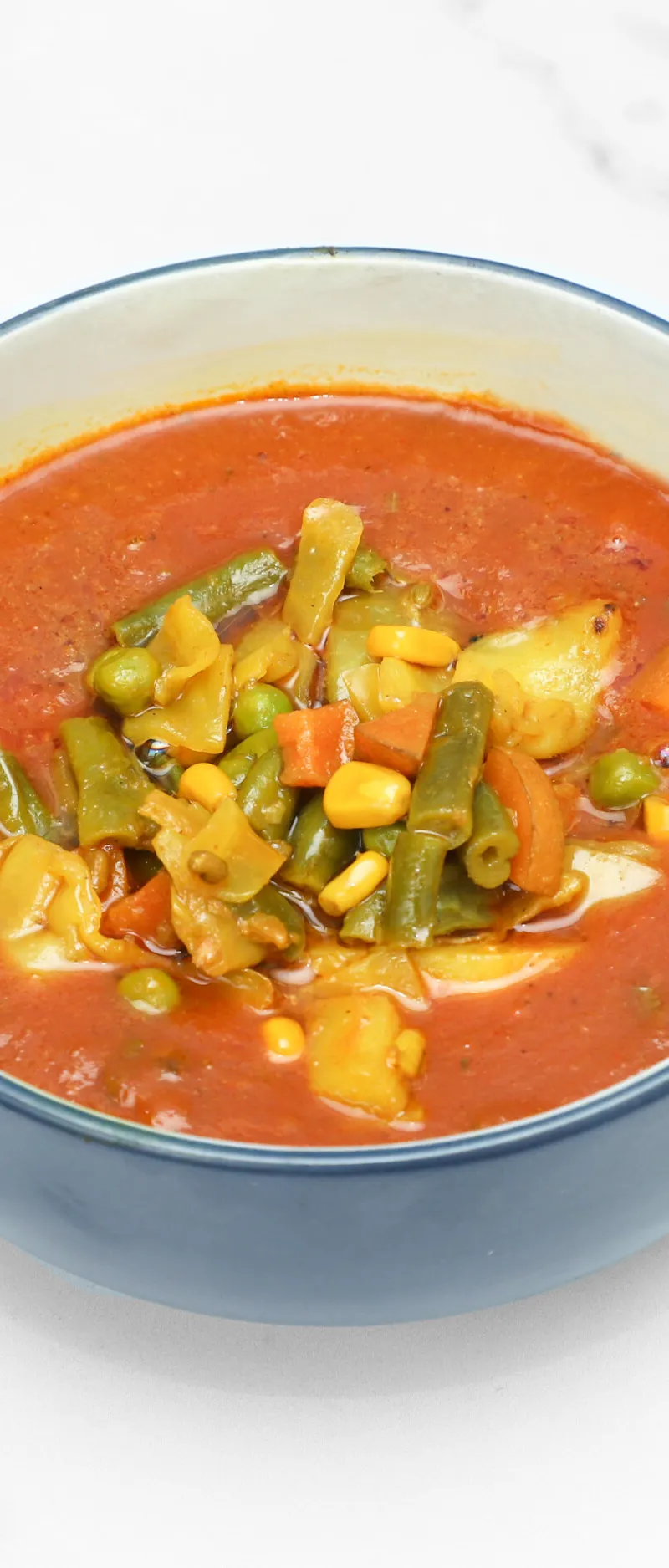 A close-up of mixed vegetable curry in a blue ceramic bowl on a white marble surface.