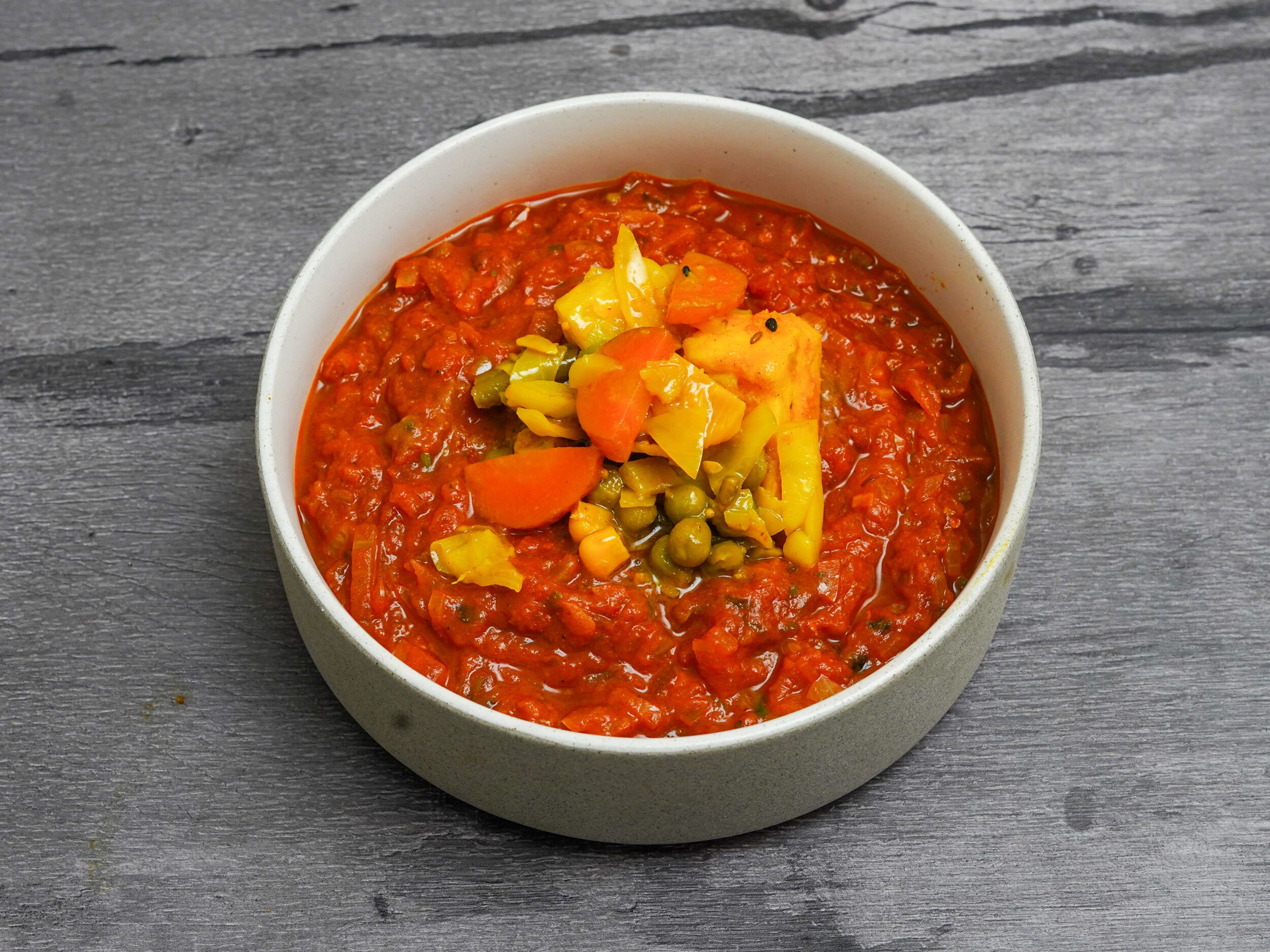 A white bowl filled with a red vegetable curry, topped with diced carrots, green peas, and yellow bell peppers.