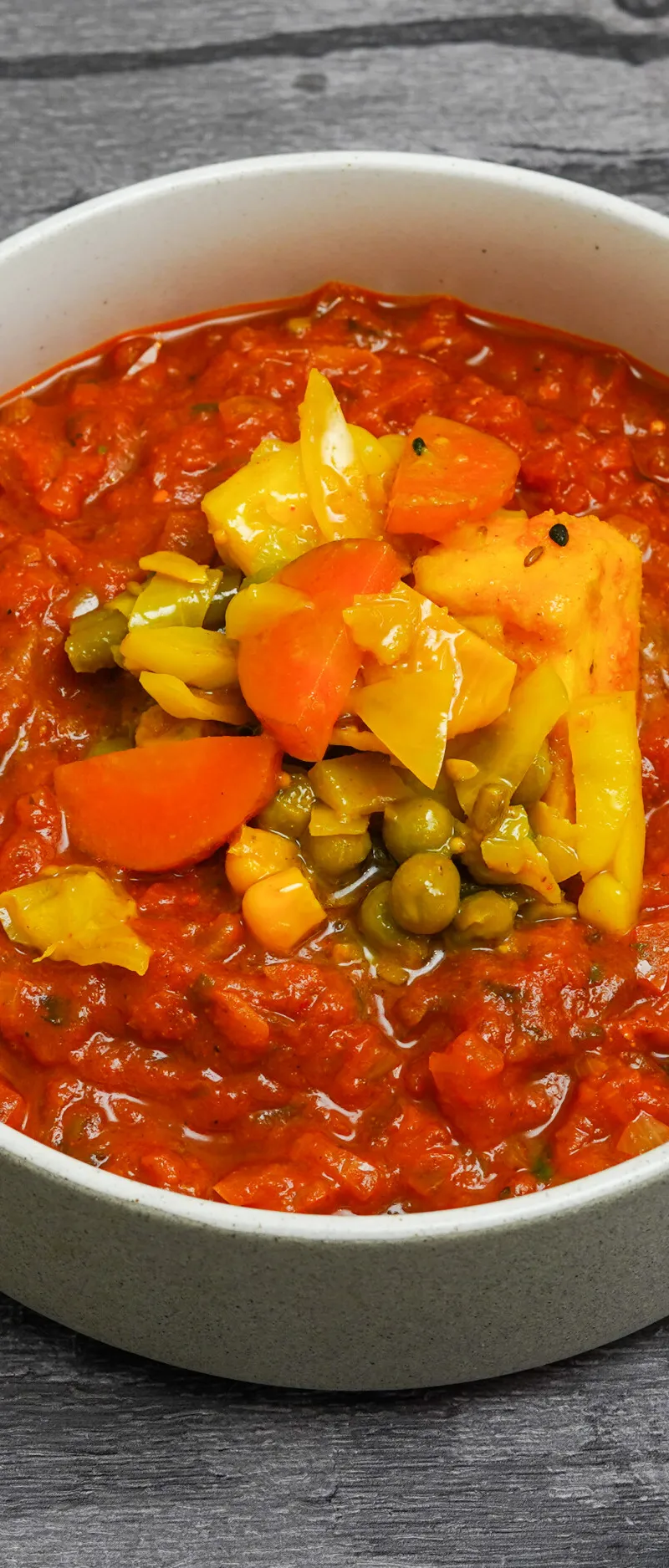 A white bowl filled with a red vegetable curry, topped with diced carrots, green peas, and yellow bell peppers.