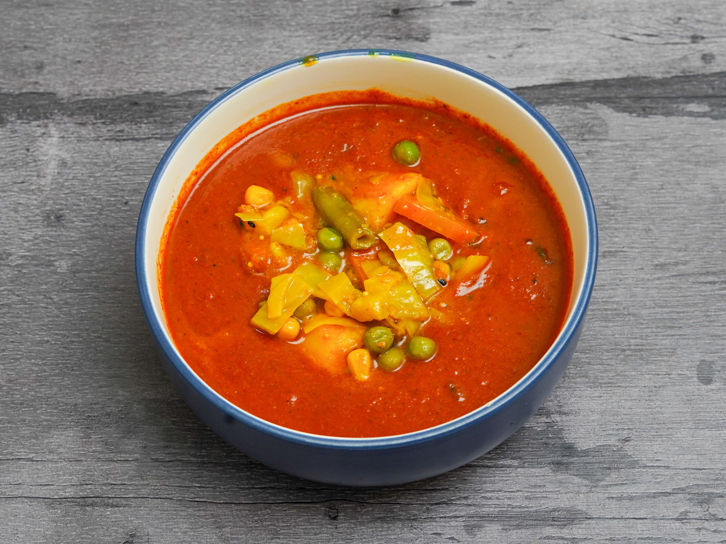 A close-up of a colorful vegetable curry in a blue bowl on a textured grey surface.