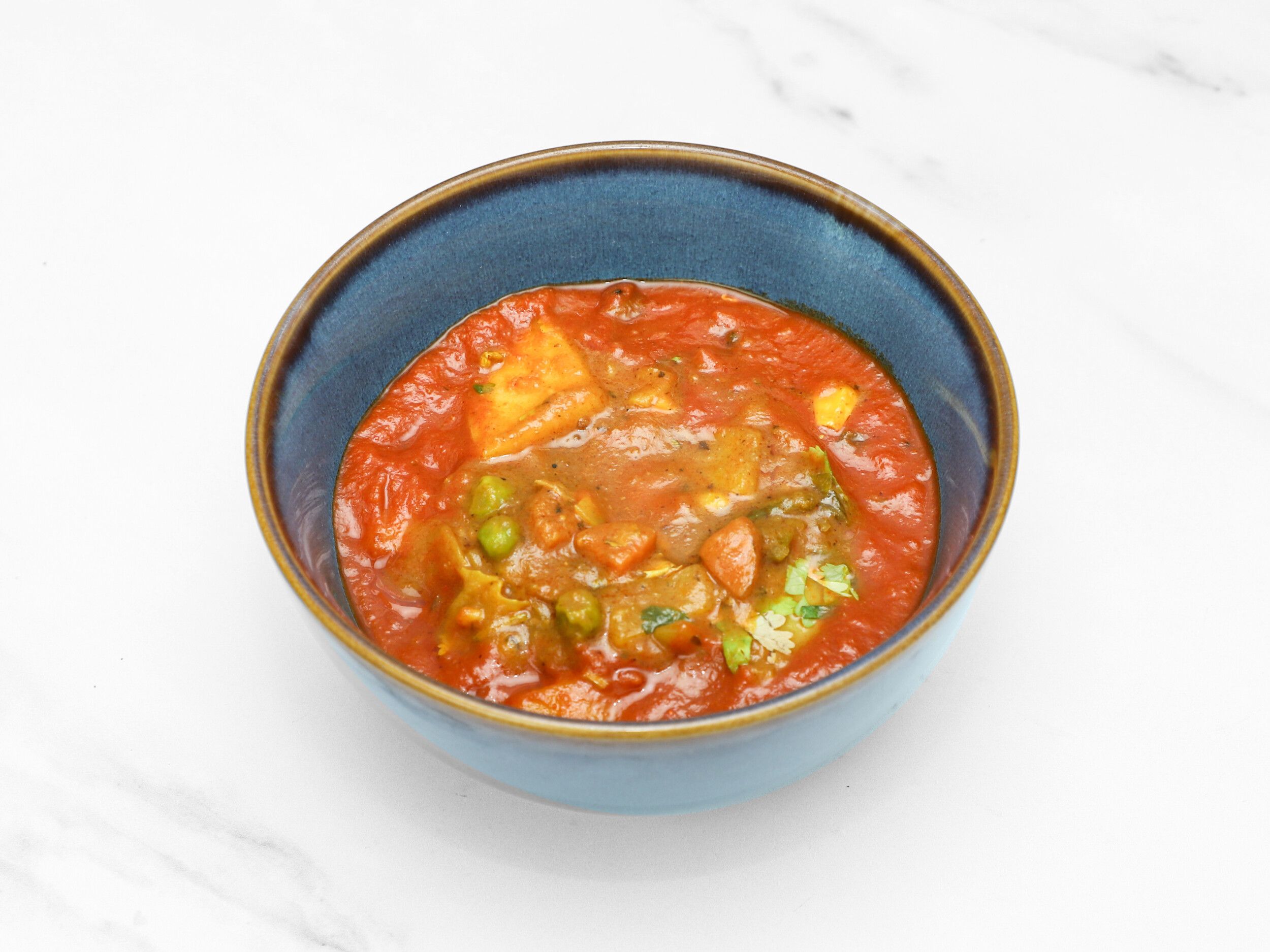 A blue ceramic bowl filled with a rich orange-red curry, featuring paneer, peas, and other vegetables. The bowl sits on a white marble surface.