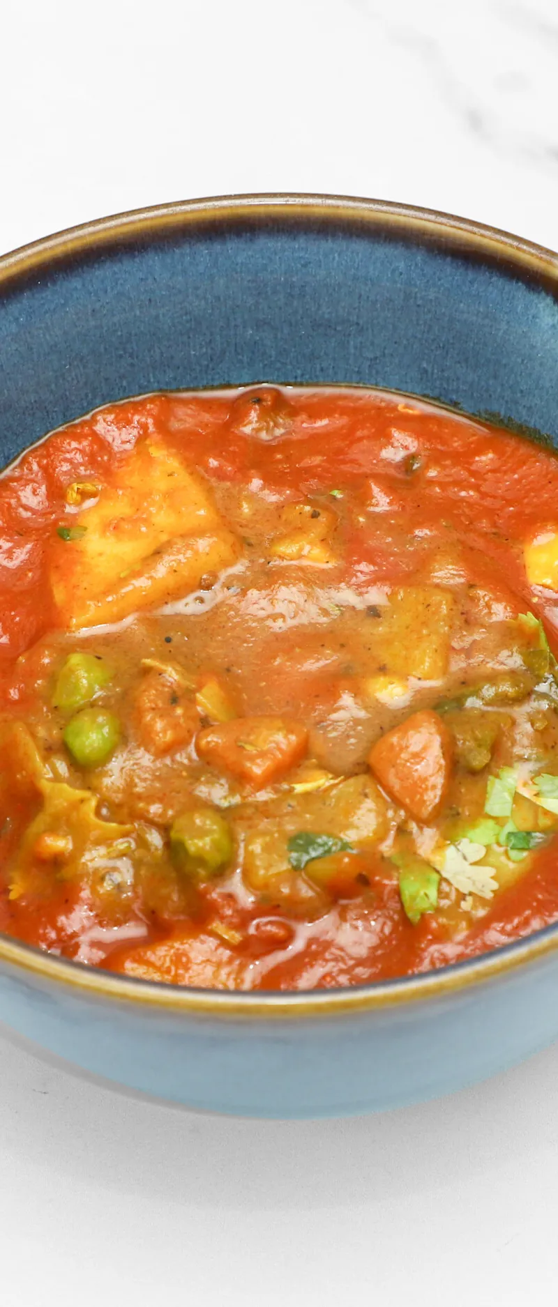 A blue ceramic bowl filled with a rich orange-red curry, featuring paneer, peas, and other vegetables. The bowl sits on a white marble surface.