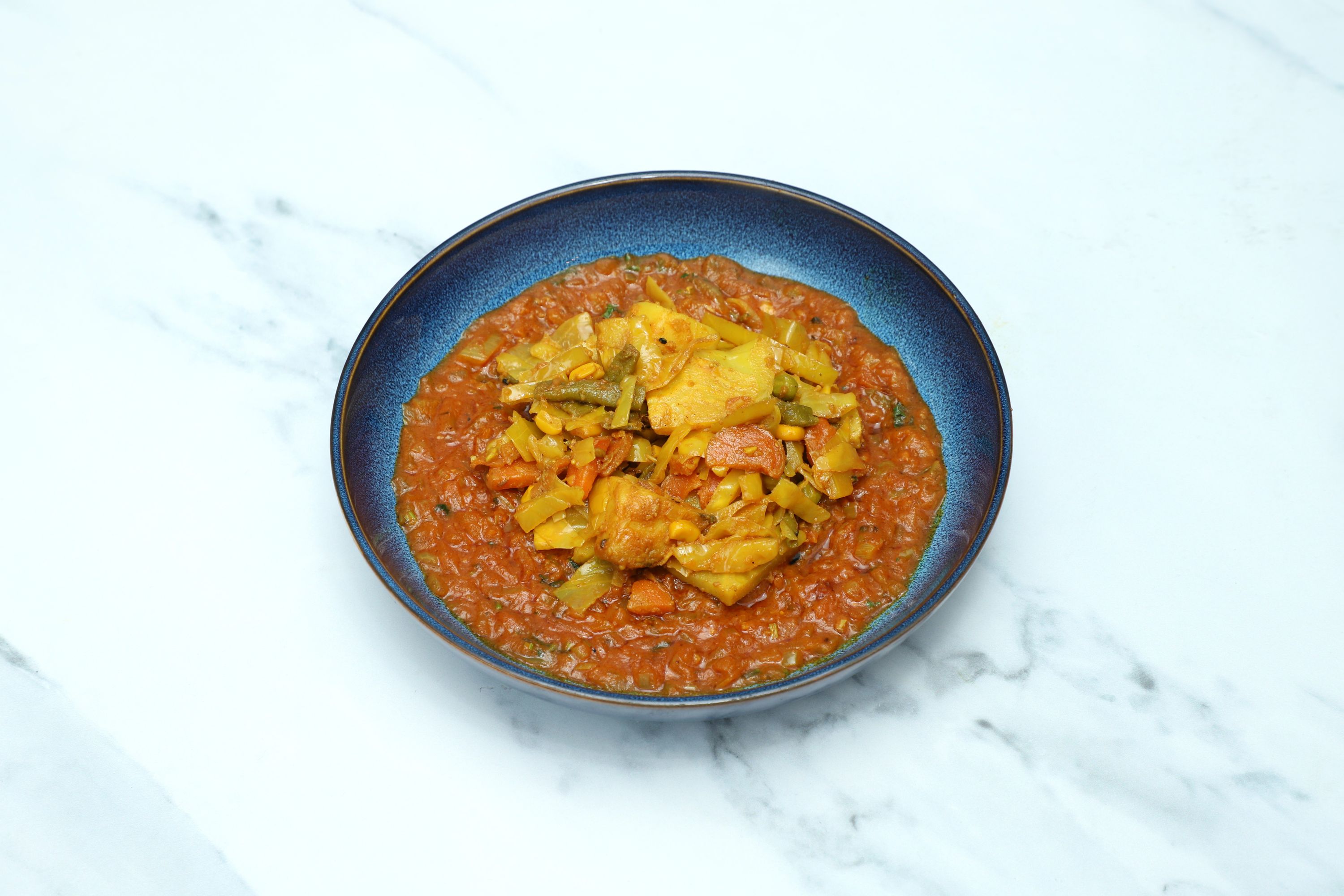 A close-up view of a vibrant vegetable curry in a blue ceramic bowl on a white marble surface.