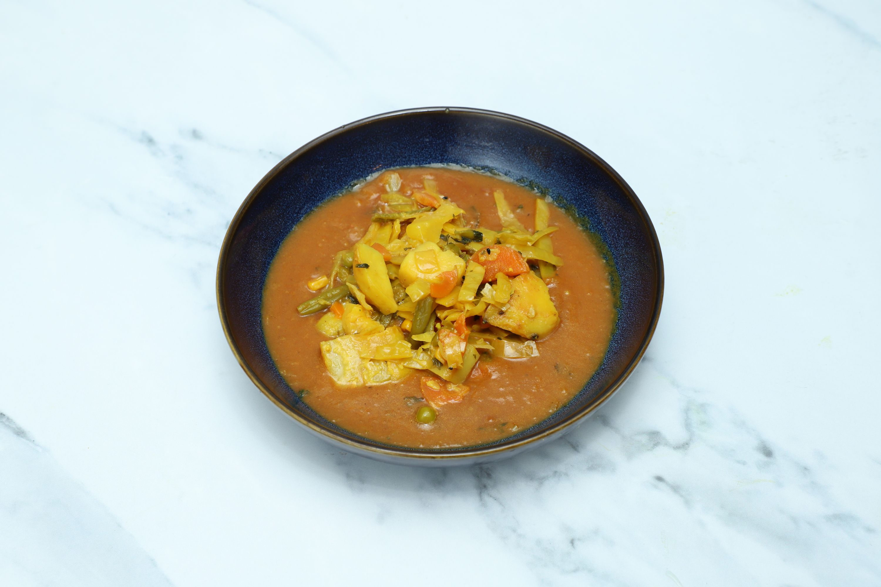 Vegetable curry with potatoes, cabbage, carrots, and green beans in a dark blue bowl on white marble.