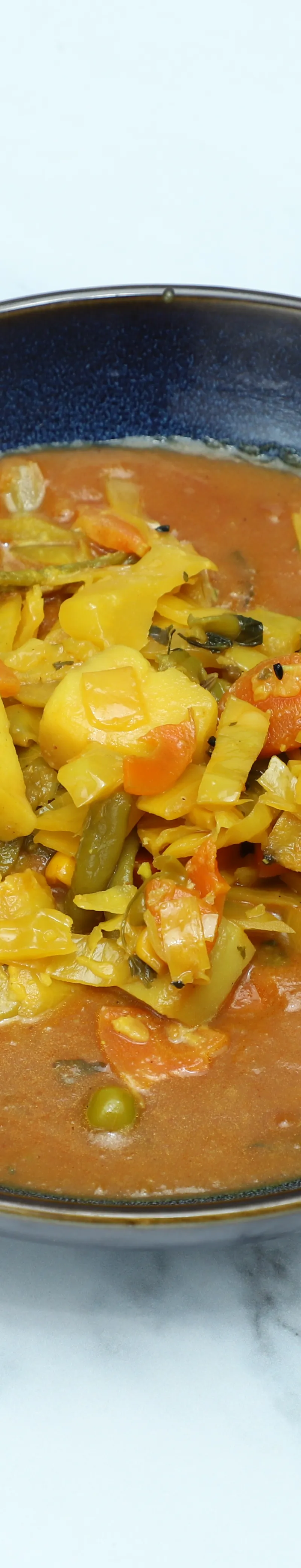 Vegetable curry with potatoes, cabbage, carrots, and green beans in a dark blue bowl on white marble.