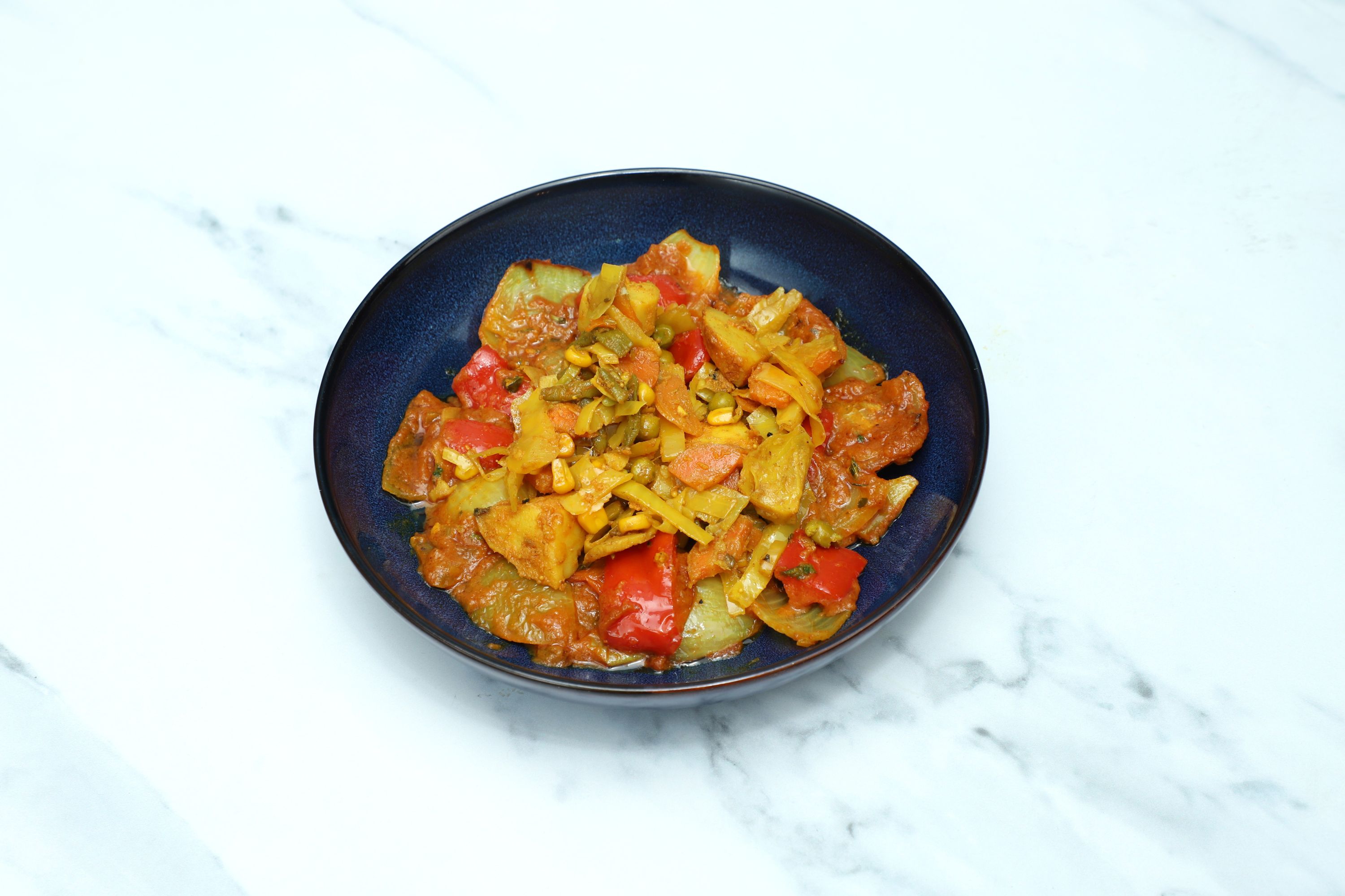 A dark blue bowl filled with a colorful vegetable bhaji on a white marble surface