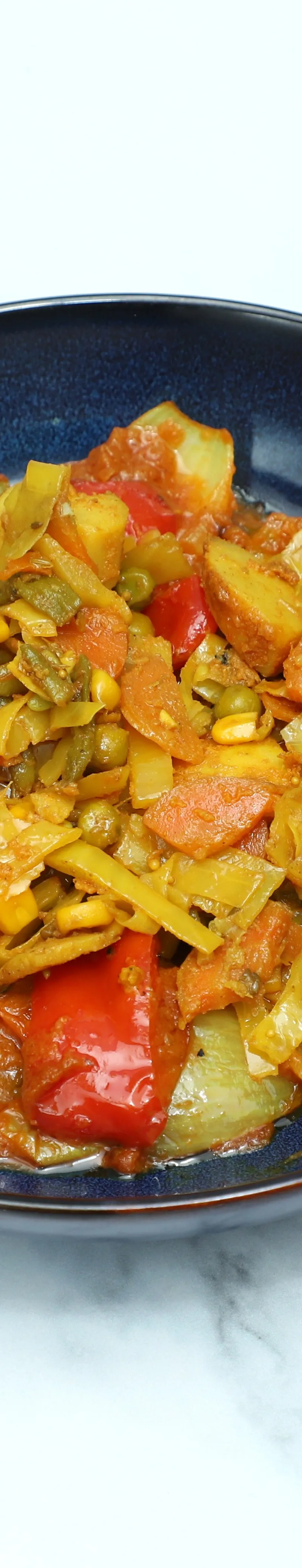 A dark blue bowl filled with a colorful vegetable bhaji on a white marble surface