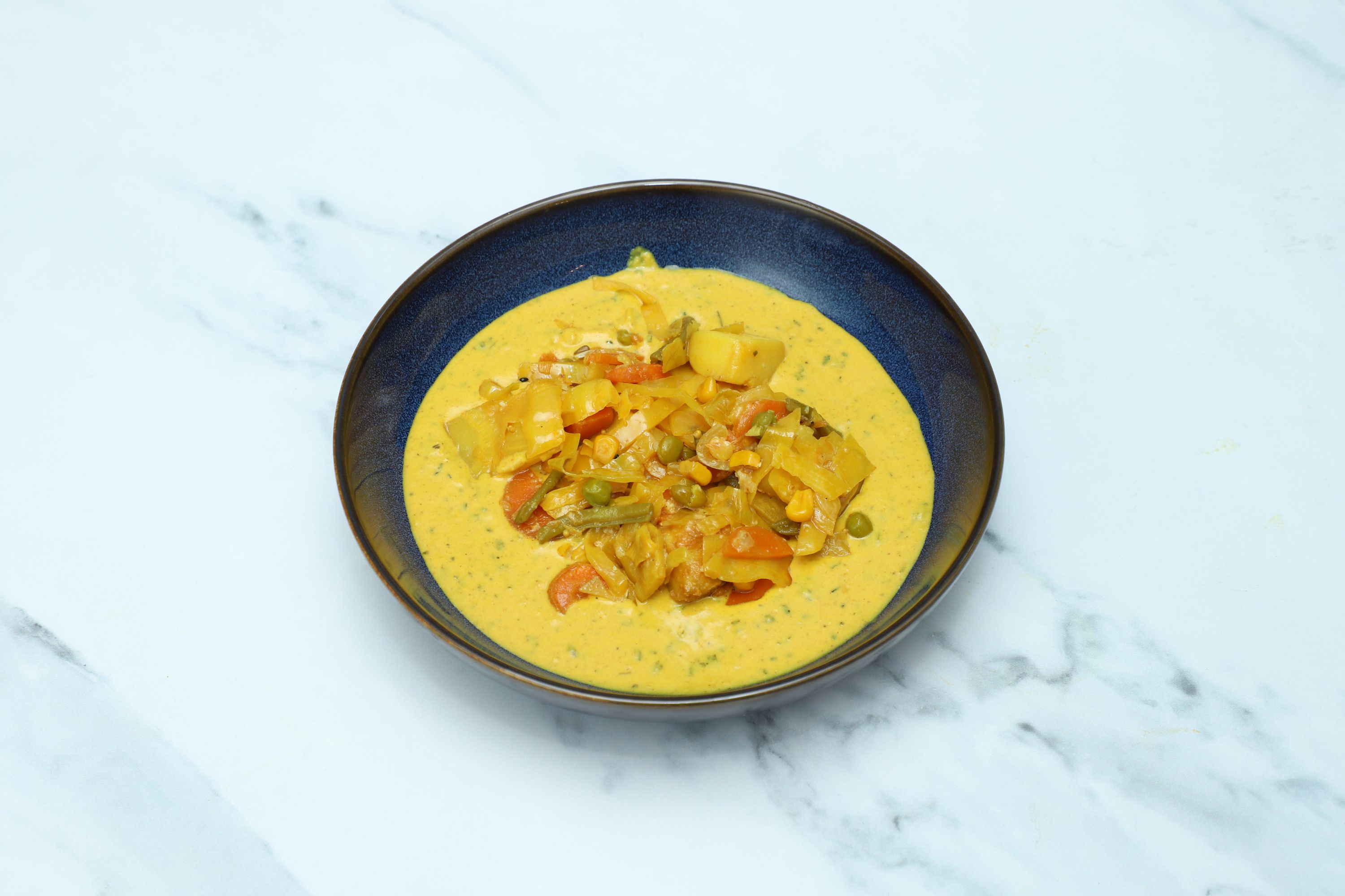 A dark blue bowl filled with a creamy yellow vegetable curry, showcasing visible carrots, peas, and potatoes.