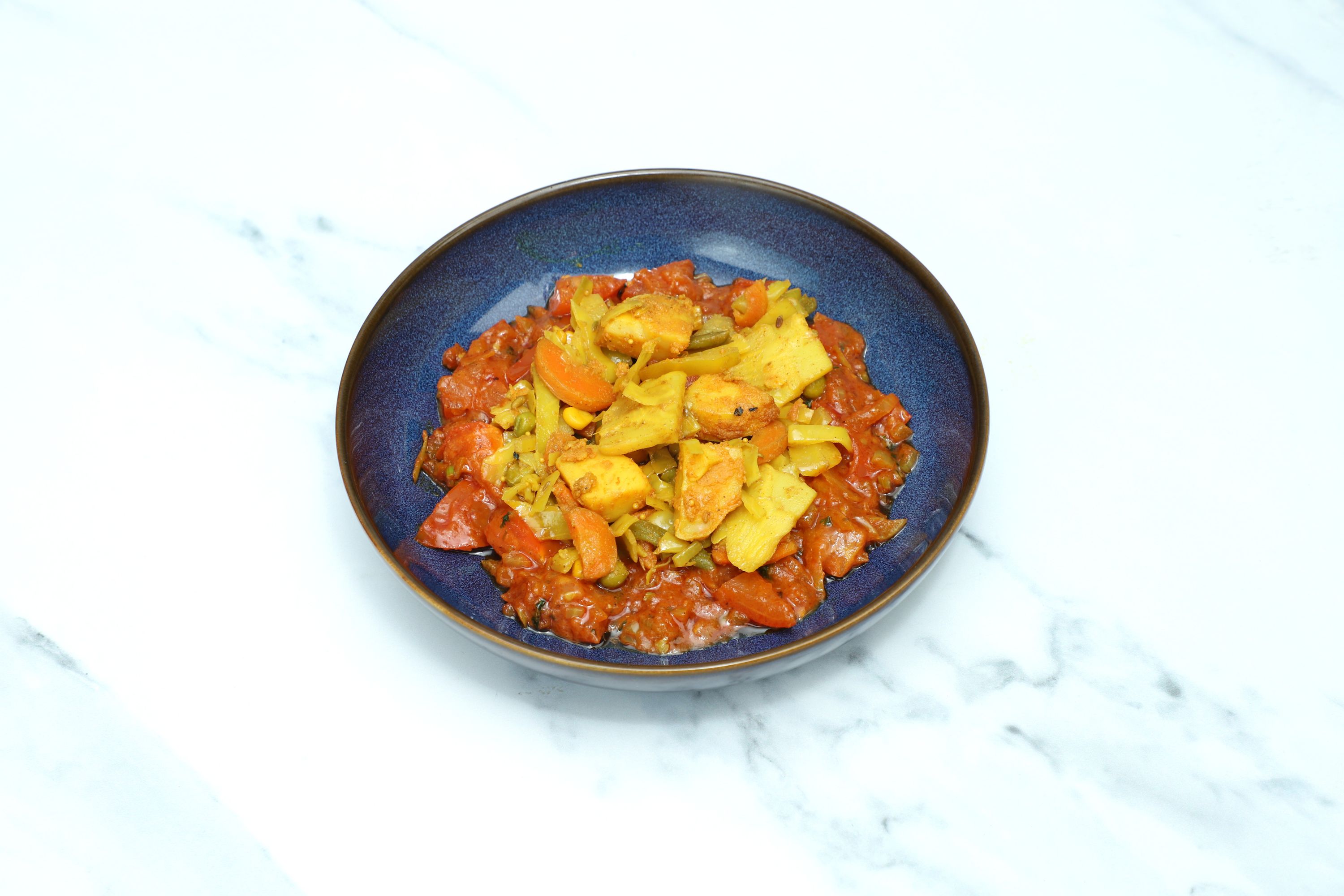A round, blue bowl filled with a colorful mix of vegetables and tomato sauce on a marble surface.