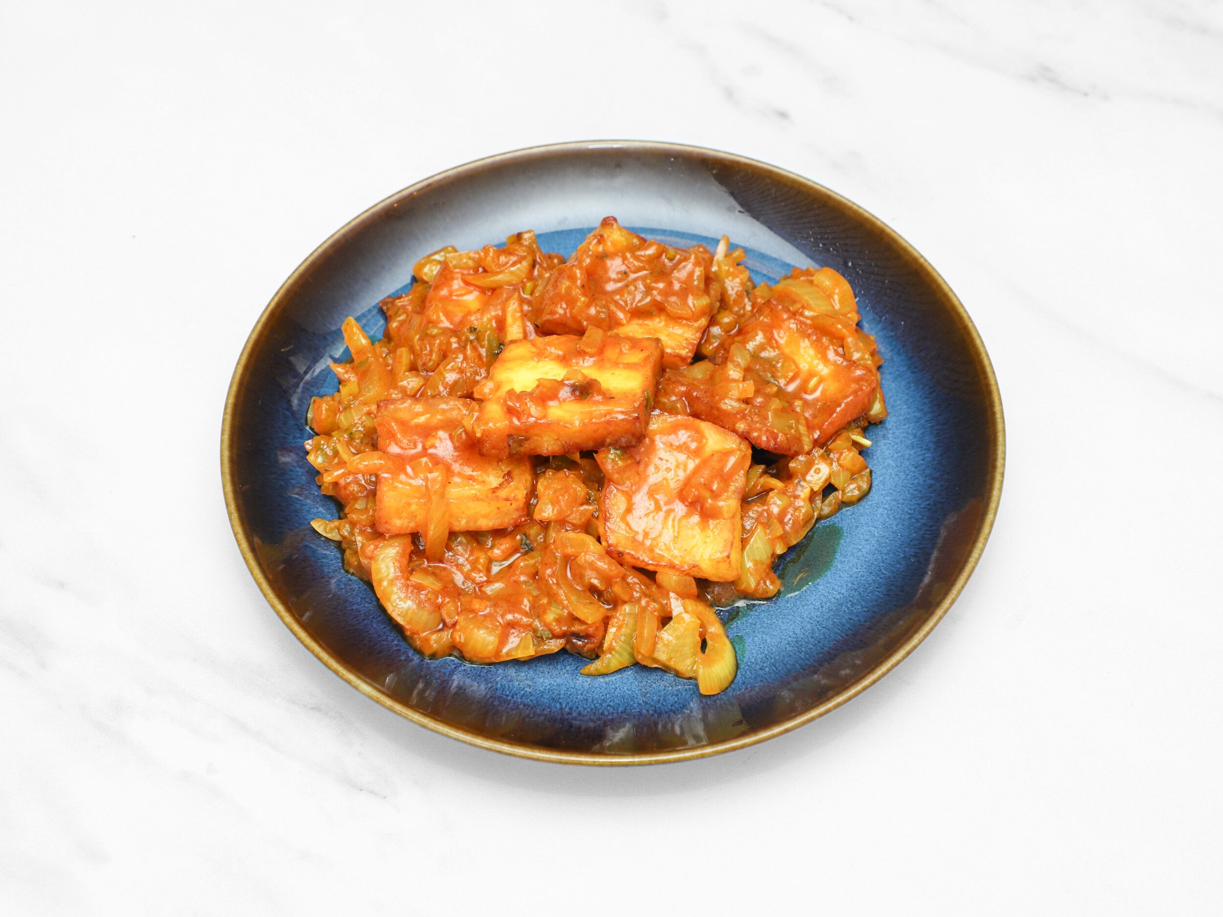Bright orange paneer chilli stir-fry on a dark blue ceramic plate against a white marble background.