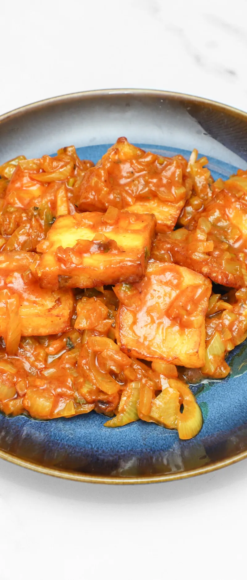 Bright orange paneer chilli stir-fry on a dark blue ceramic plate against a white marble background.