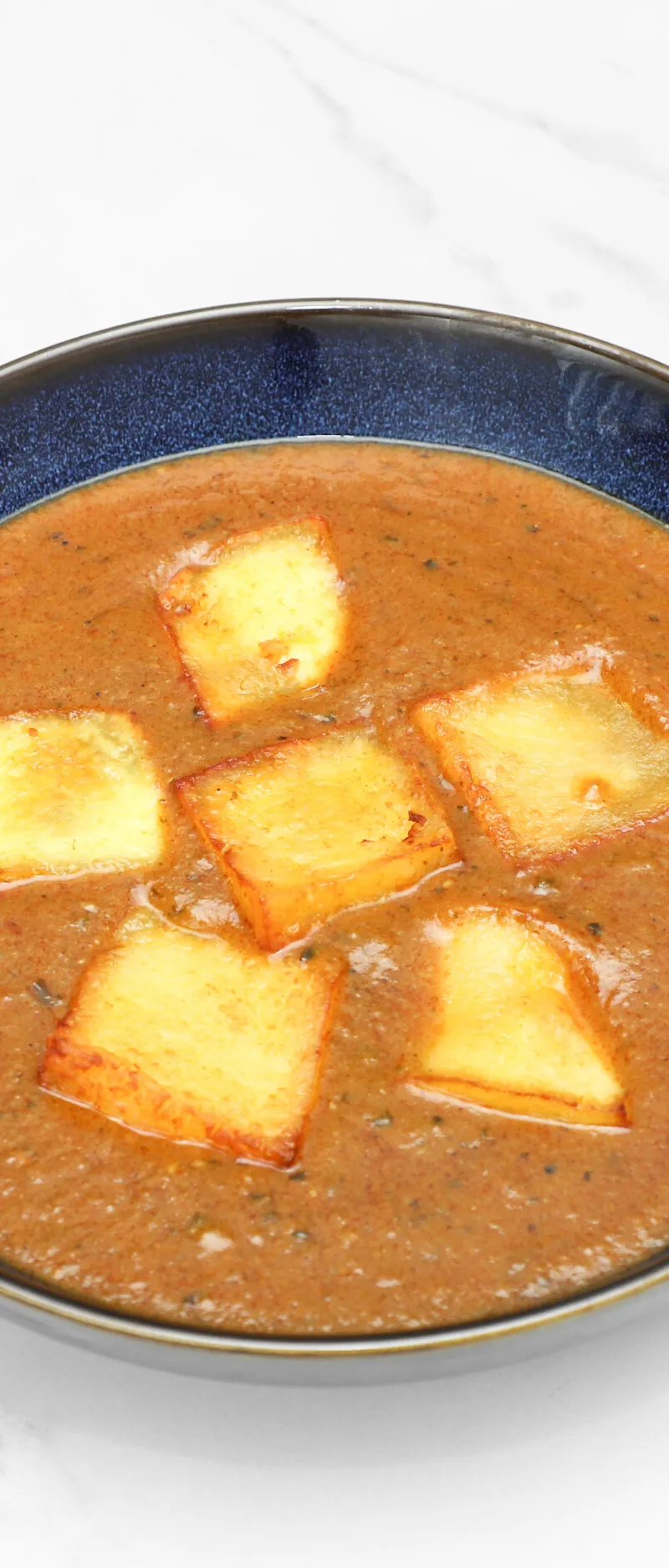 Top-down view of a dark blue bowl filled with a brown curry and five fried halloumi pieces.