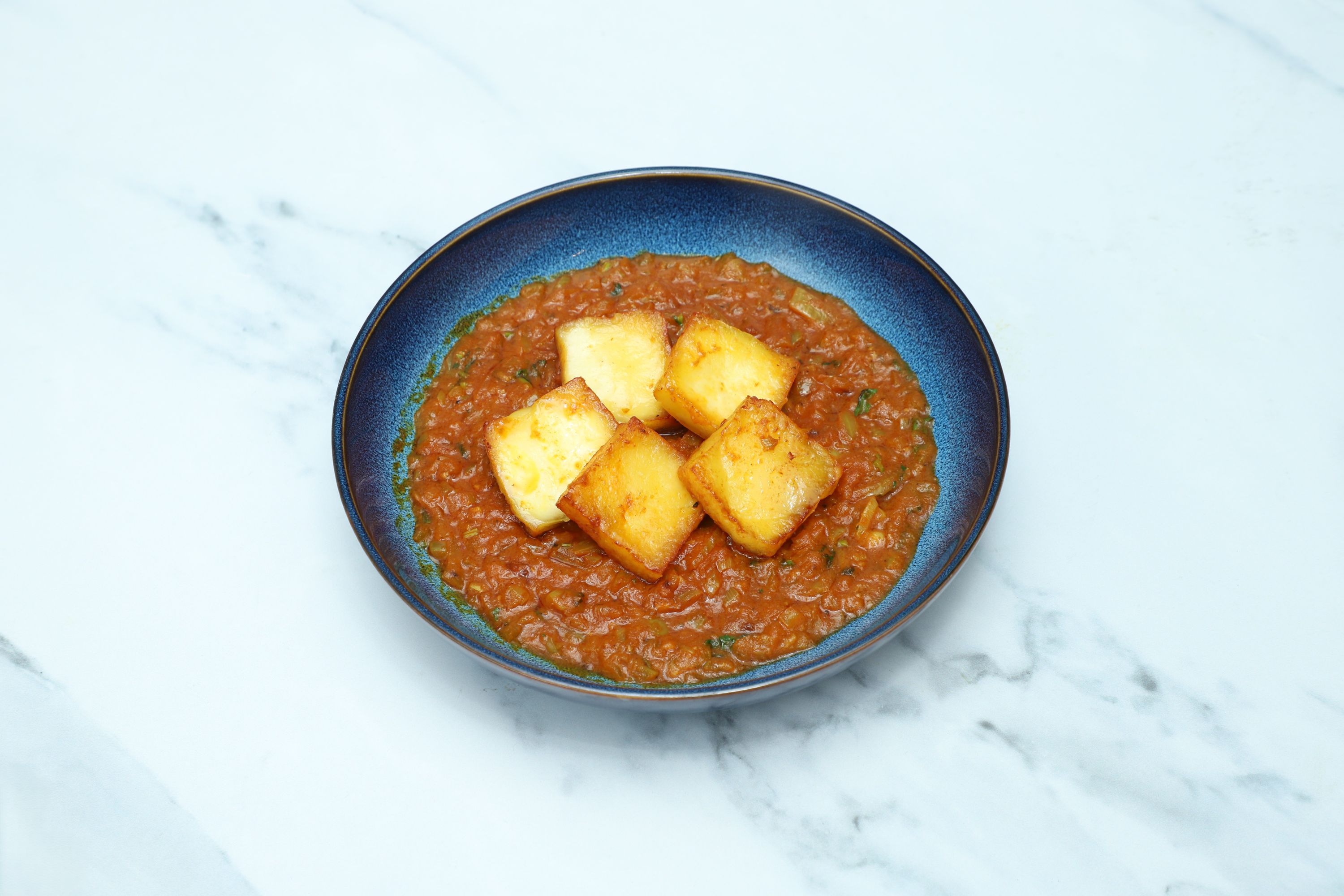 A round blue dish filled with a rustic orange-red curry and five square pieces of fried cheese
