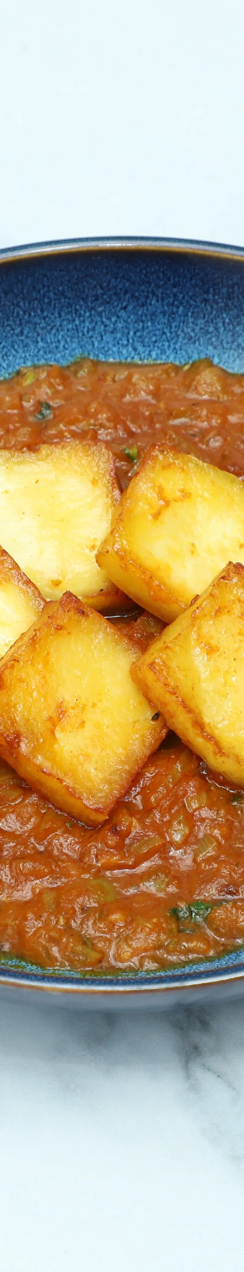 A round blue dish filled with a rustic orange-red curry and five square pieces of fried cheese