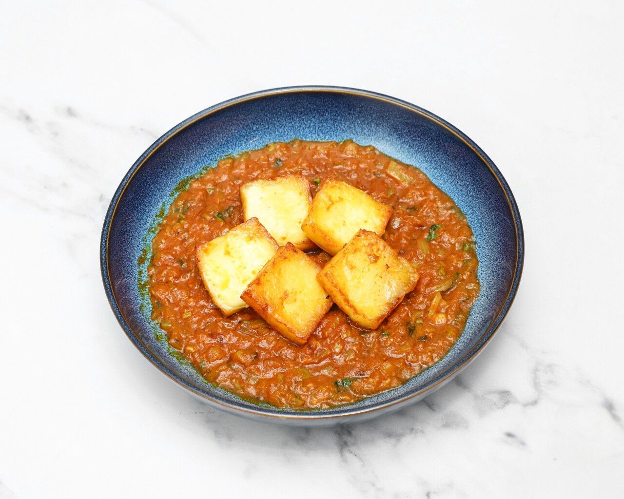 Five square pieces of golden-brown paneer cheese in a rich orange-red curry sauce in a blue ceramic bowl.
