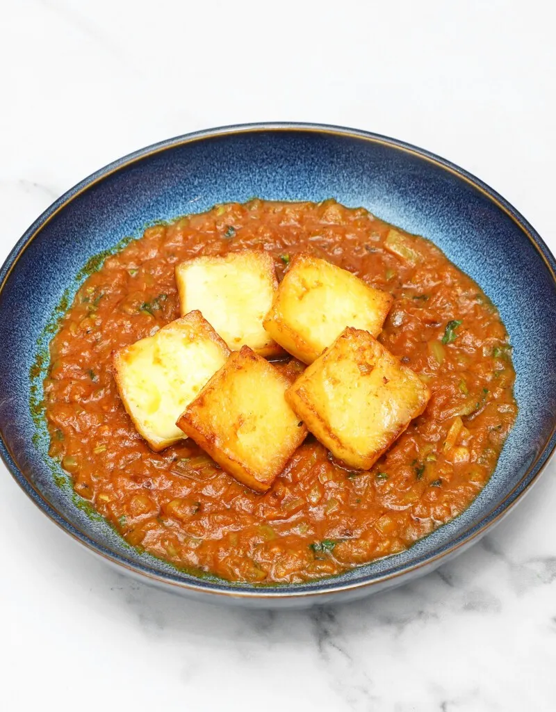 Five square pieces of golden-brown paneer cheese in a rich orange-red curry sauce in a blue ceramic bowl.