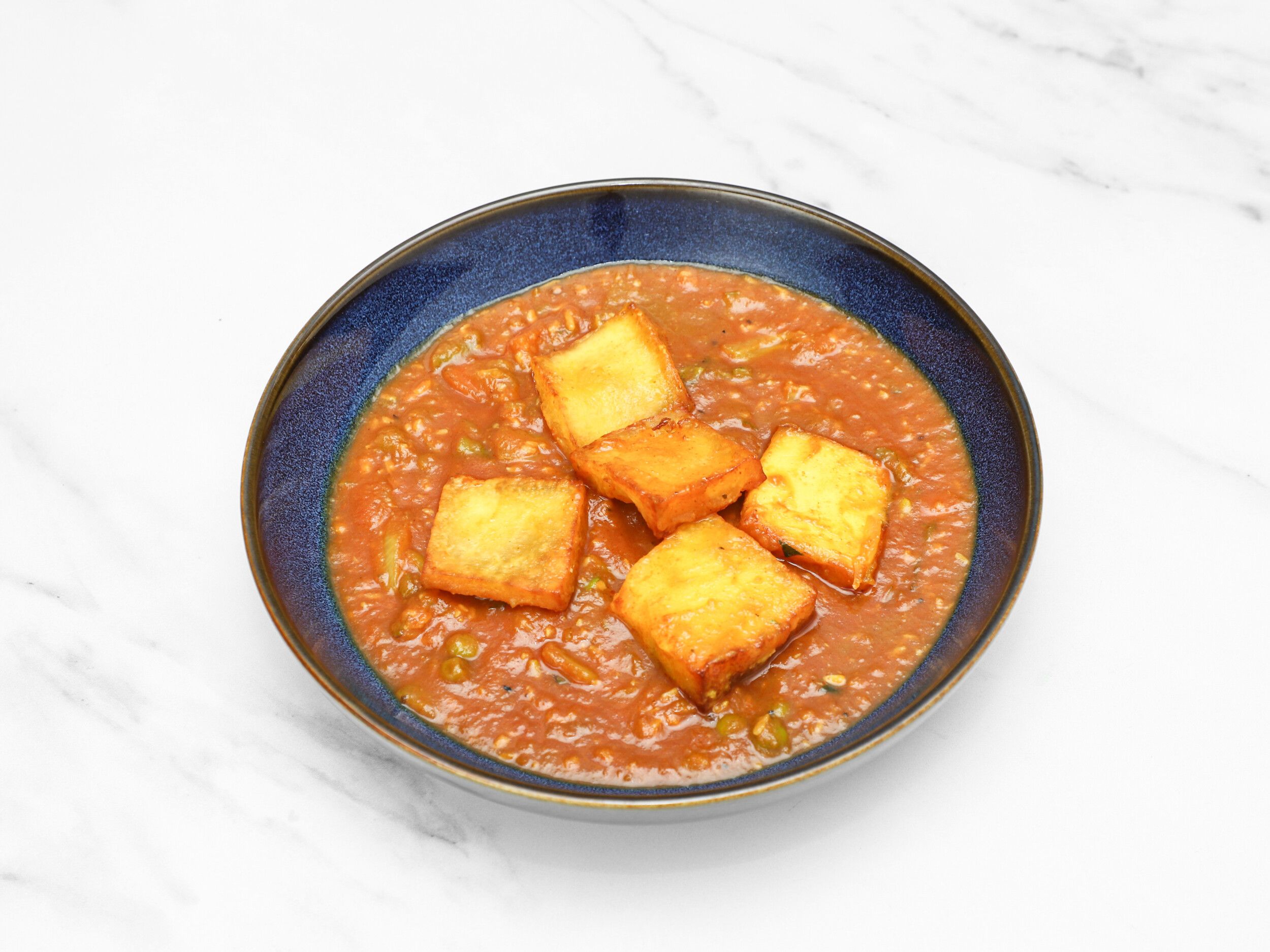 Four golden brown paneer pieces sit in a rich orange-red curry sauce in a blue bowl.