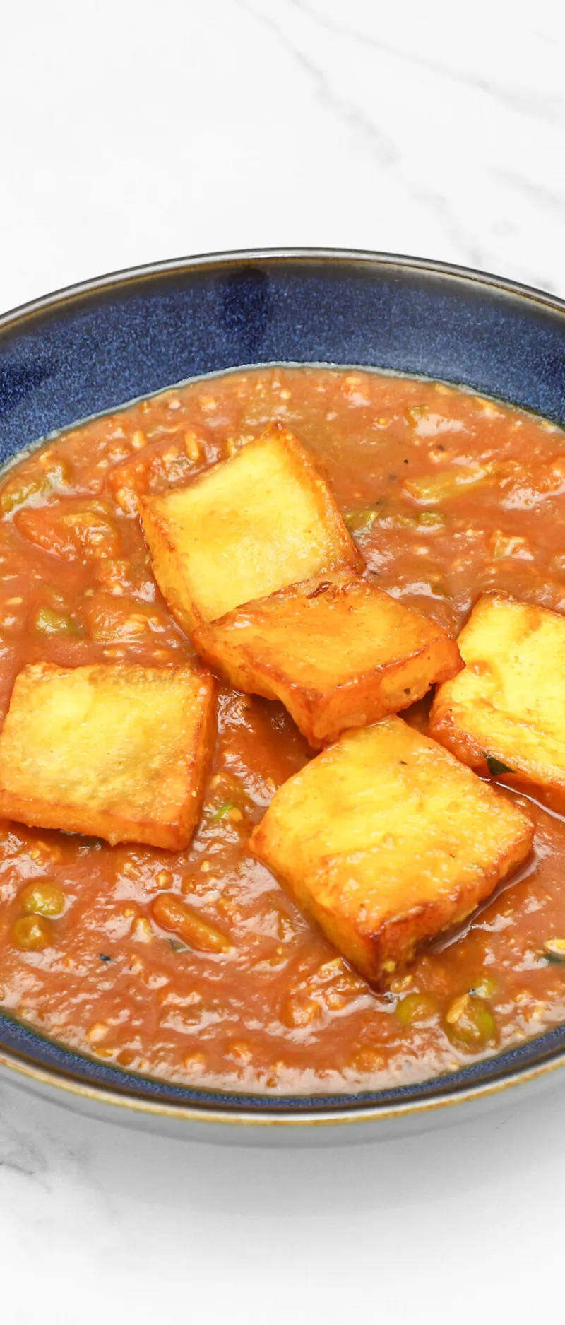 Four golden brown paneer pieces sit in a rich orange-red curry sauce in a blue bowl.