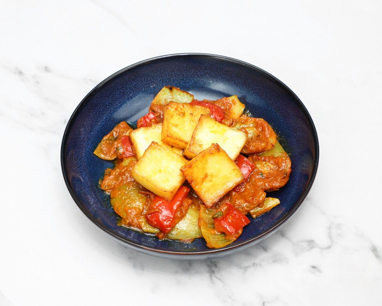 Square pieces of golden-brown paneer cheese with red and green peppers in a spicy sauce.