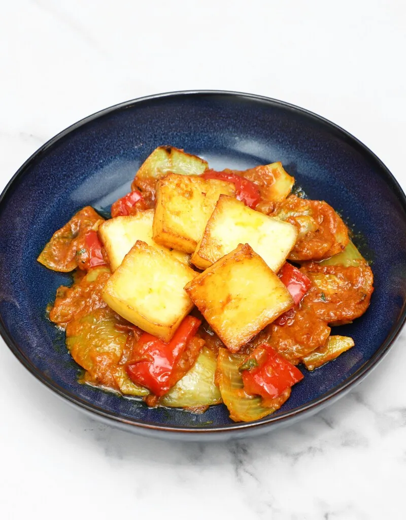 Square pieces of golden-brown paneer cheese with red and green peppers in a spicy sauce.