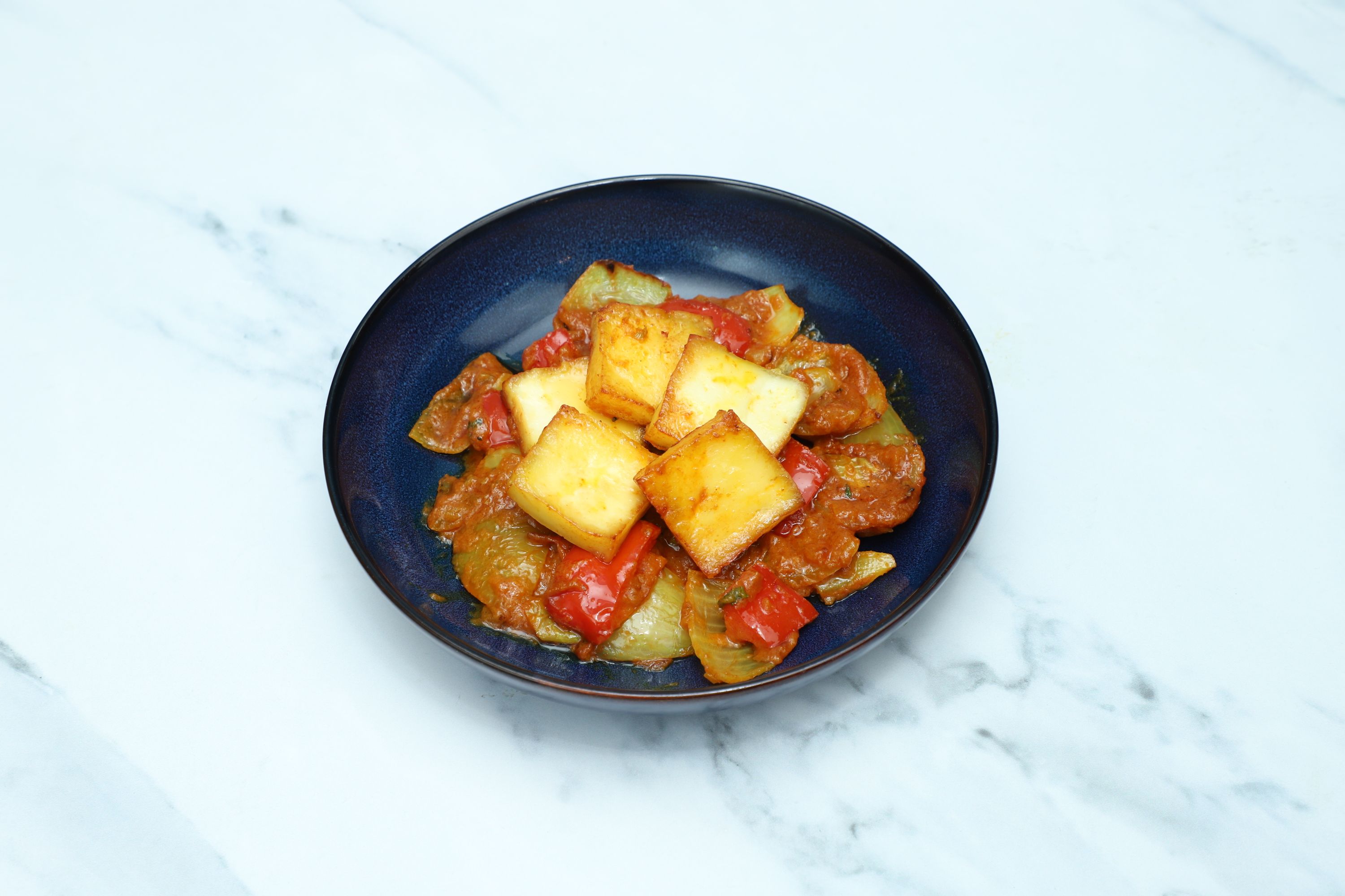 Golden paneer cubes with green and red bell peppers in a rich orange sauce, served in a deep blue bowl.