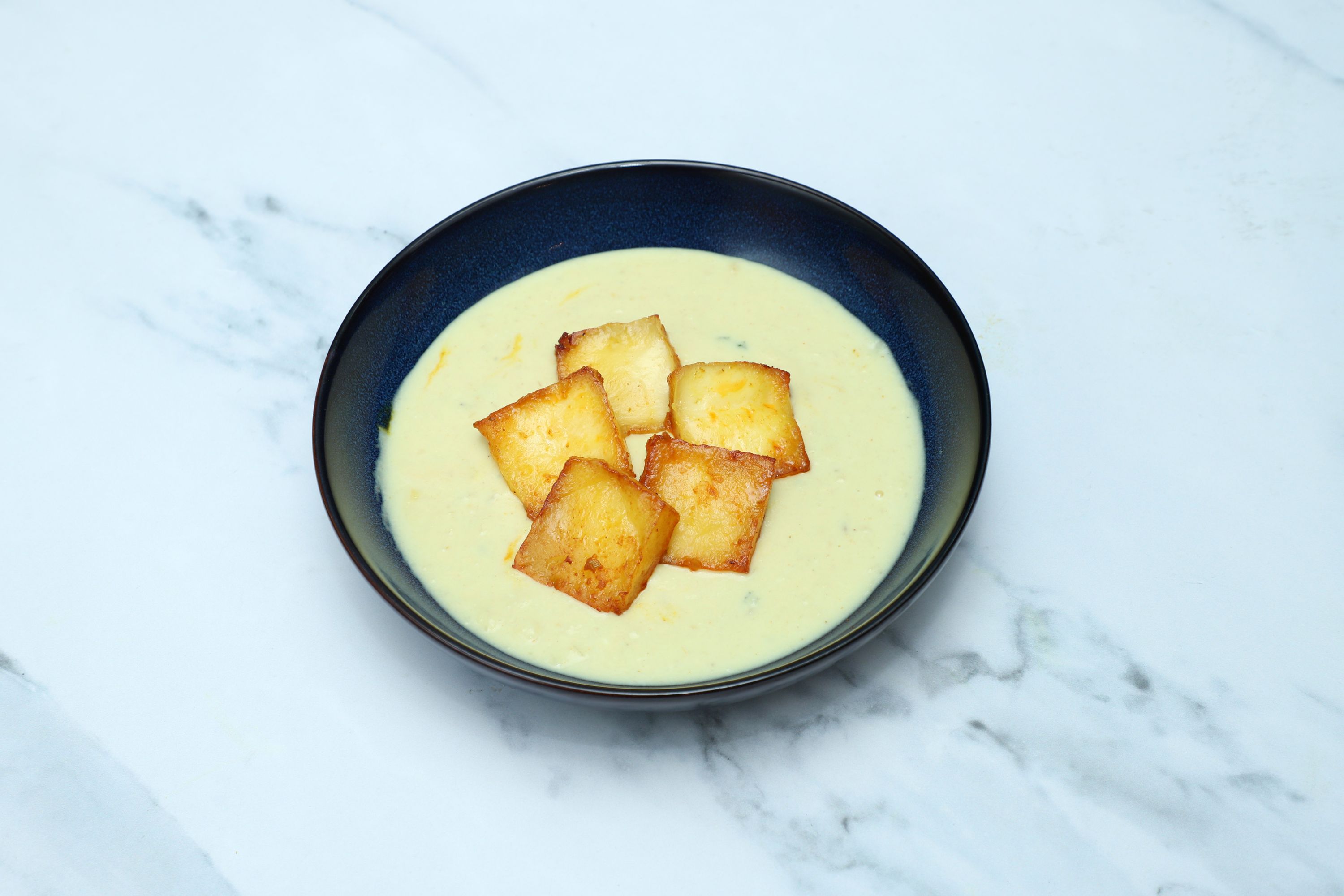 Creamy pale yellow soup in a dark bowl, topped with five golden pan-fried halloumi cubes.