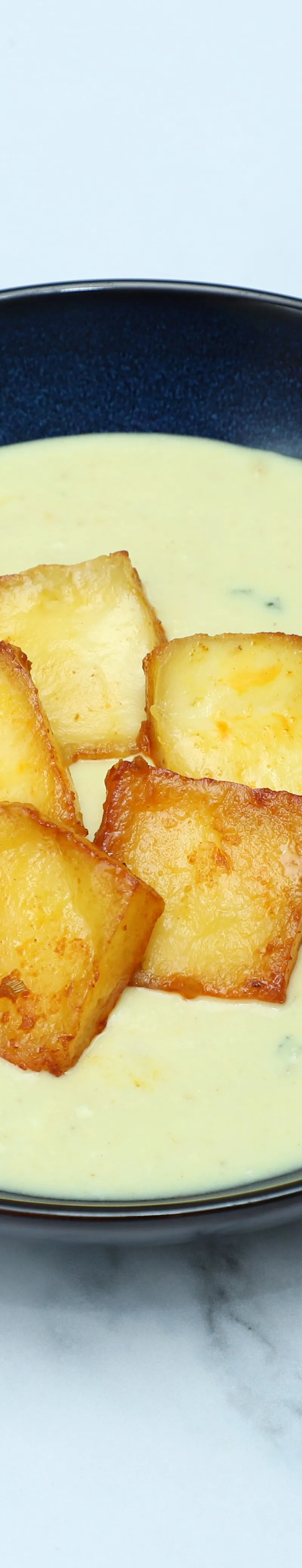 Creamy pale yellow soup in a dark bowl, topped with five golden pan-fried halloumi cubes.