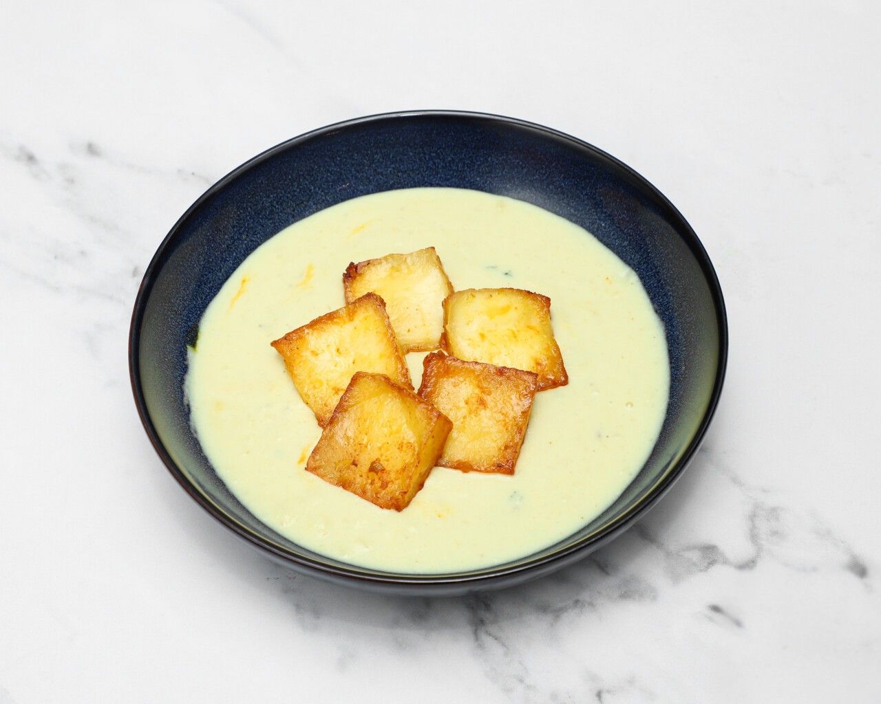 Pale yellow creamy soup with five golden-brown fried halloumi squares in a dark blue bowl.