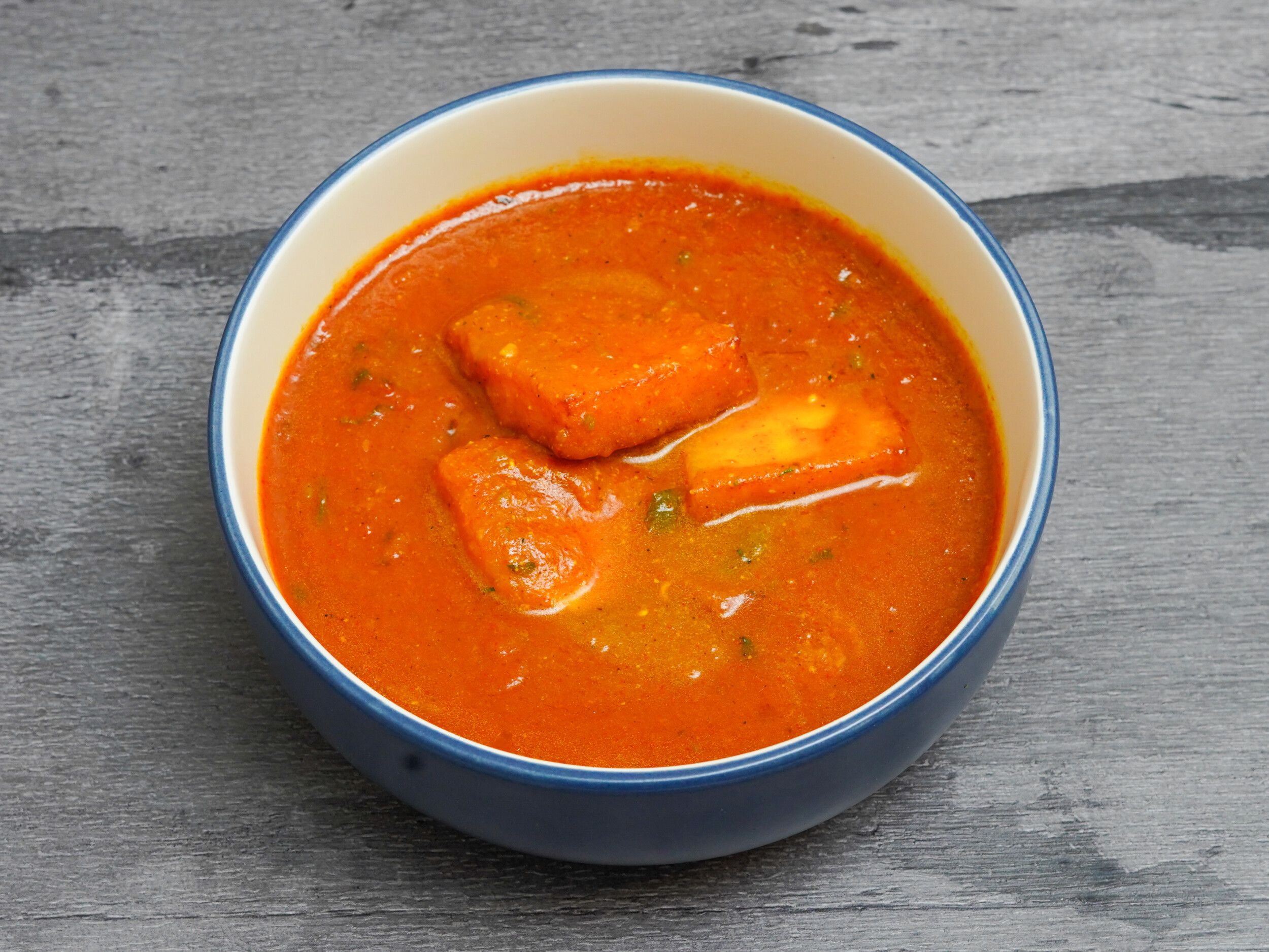 Bright orange paneer curry served in a blue bowl with a white rim on a wooden surface.