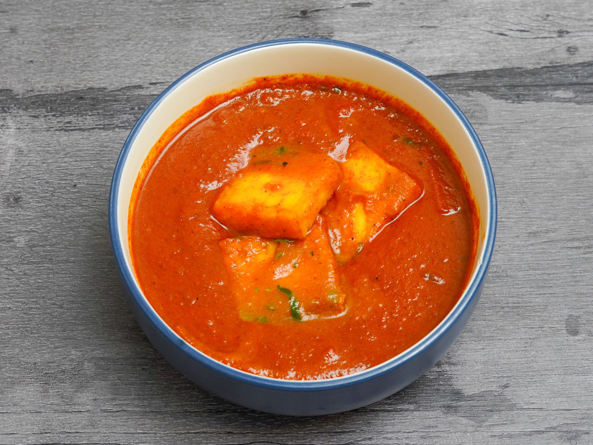 Orange paneer butter masala with three large paneer cubes in a blue and white bowl