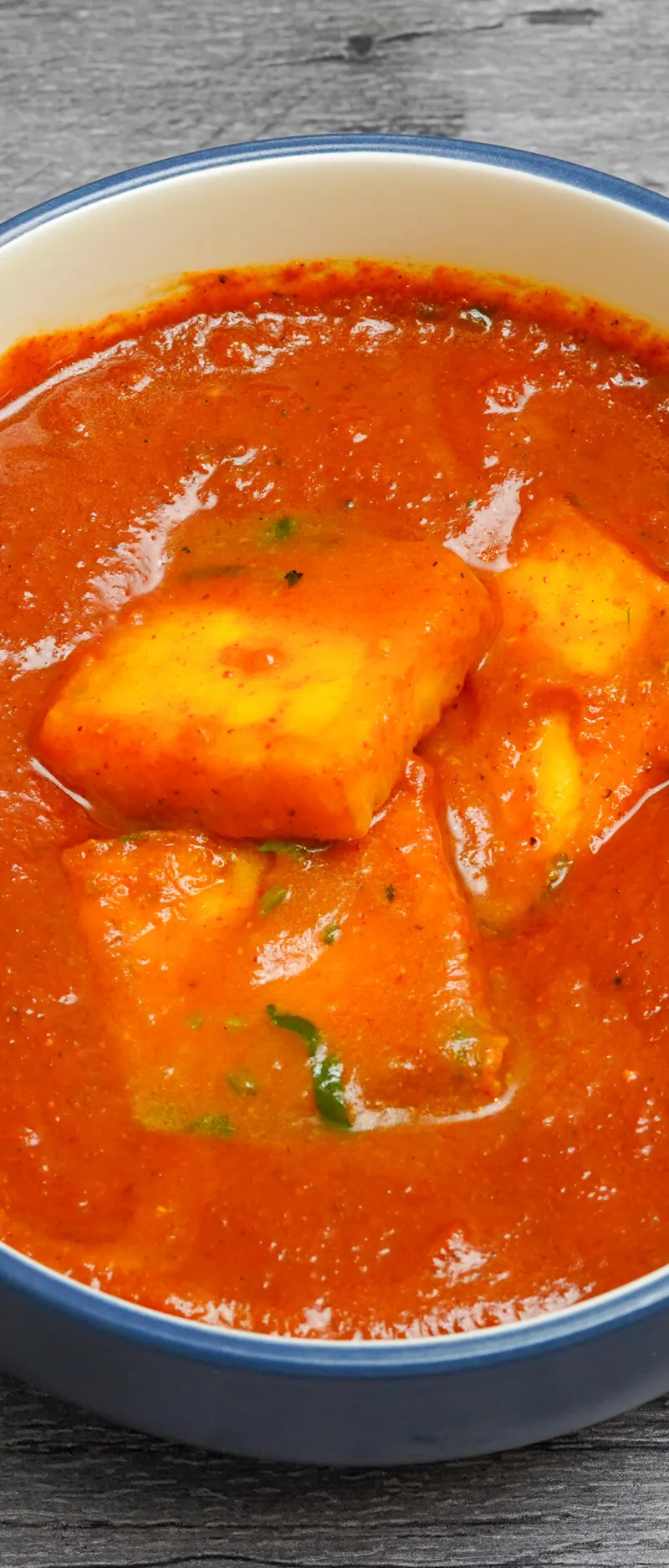 Orange paneer butter masala with three large paneer cubes in a blue and white bowl
