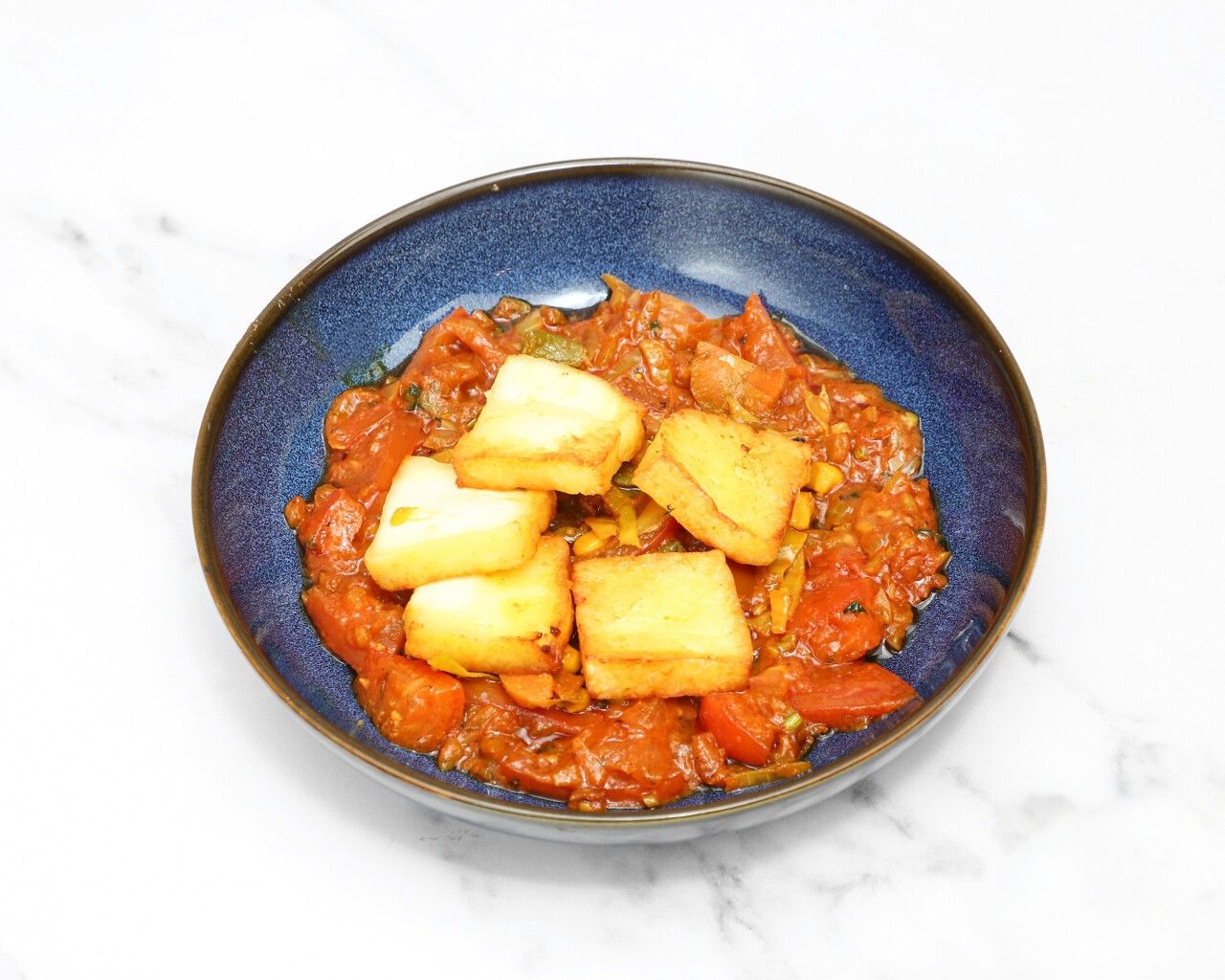Golden-brown fried paneer cubes resting on a vibrant red tomato and vegetable curry in a dark blue bowl.