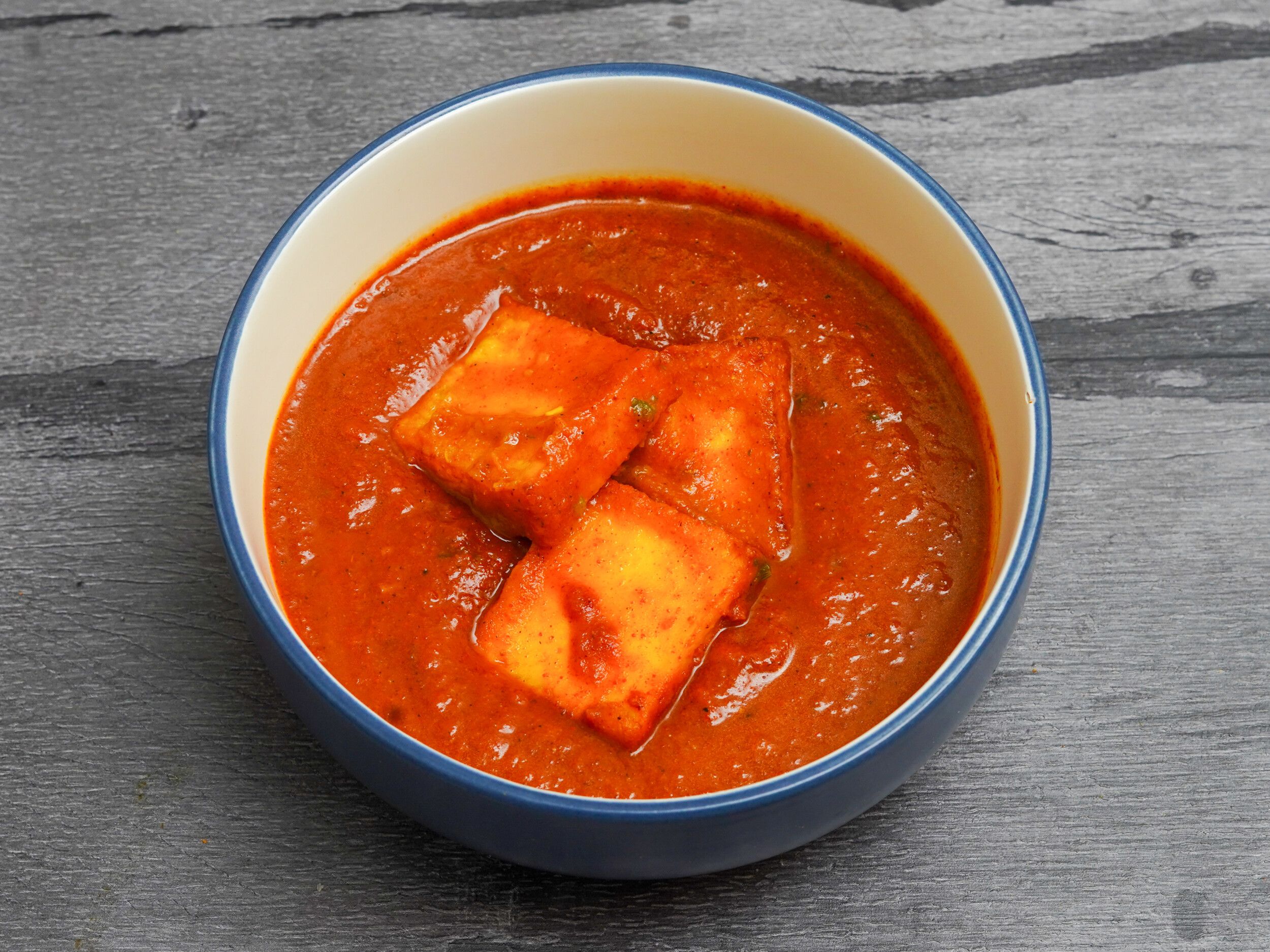 Three large chunks of paneer cheese floating in a vibrant orange-red curry sauce in a blue-rimmed bowl.