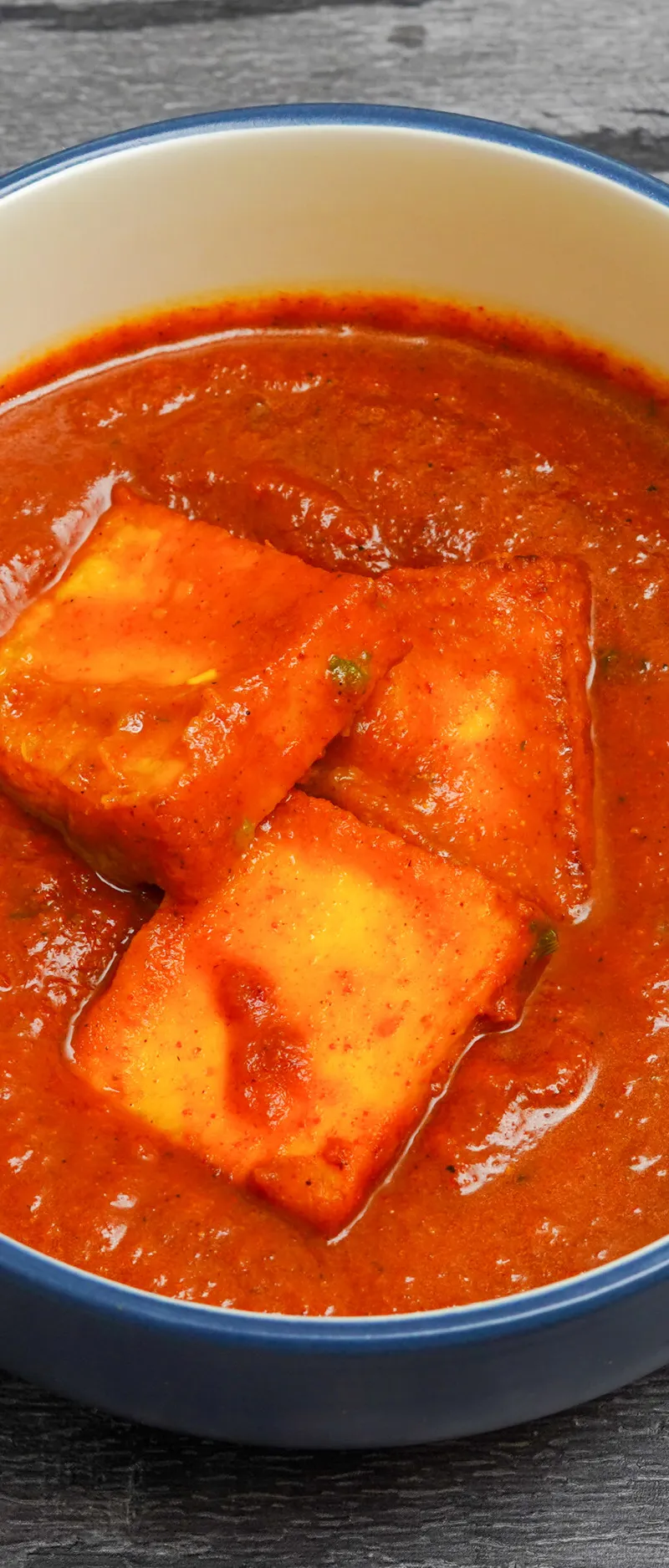 Three large chunks of paneer cheese floating in a vibrant orange-red curry sauce in a blue-rimmed bowl.