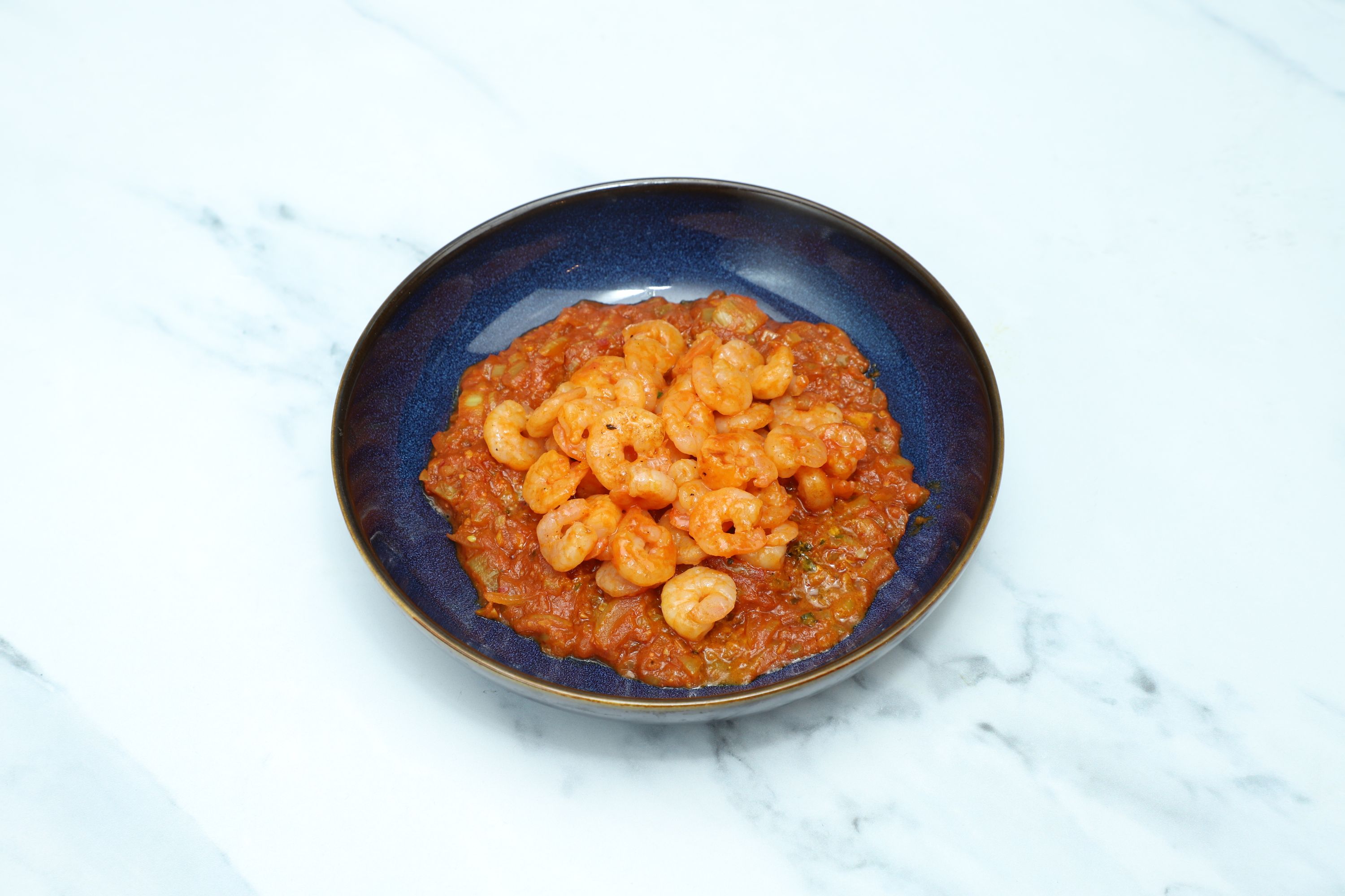 Small shrimp in a vibrant red curry sauce, served in a dark blue bowl.