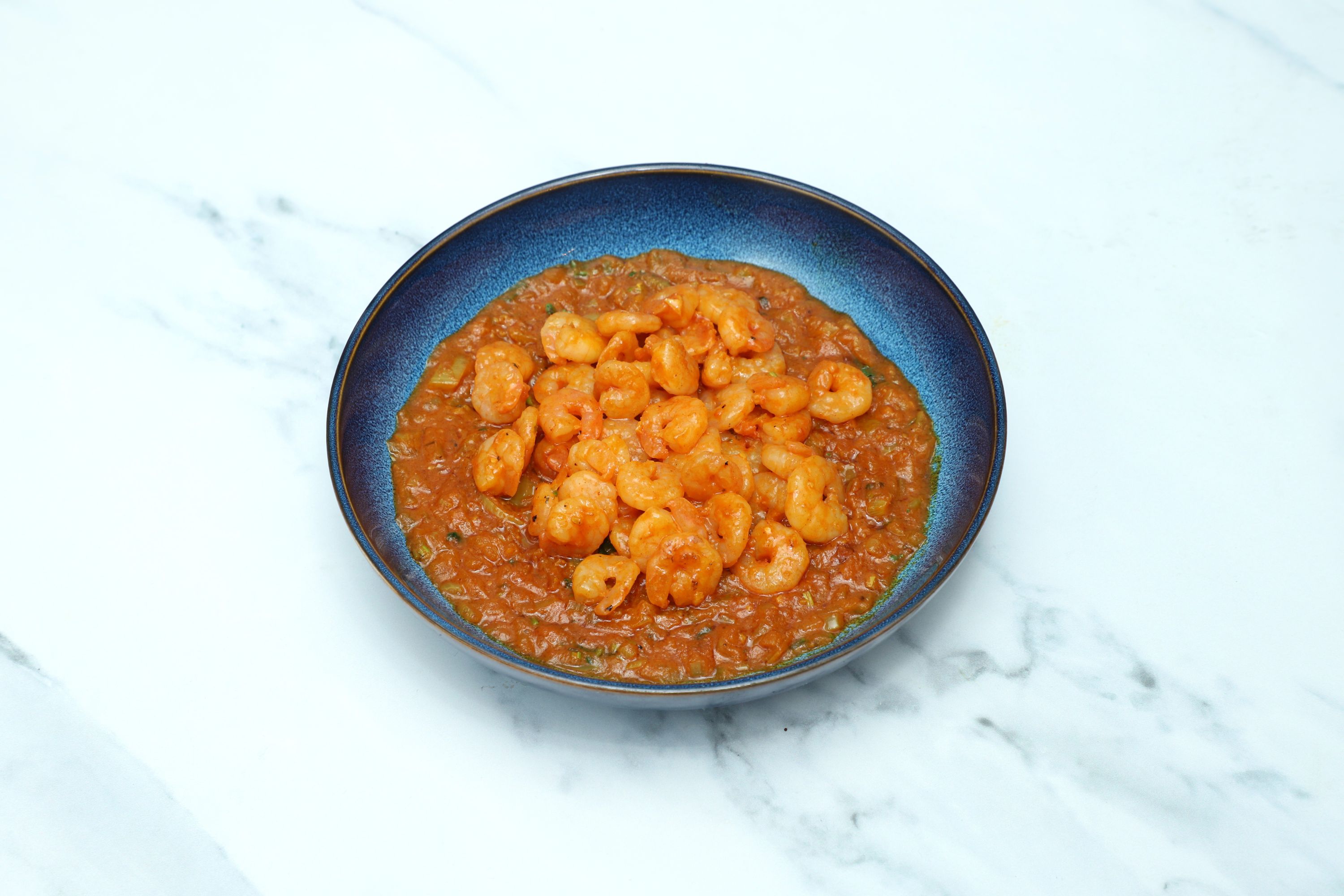 A blue bowl holds cooked king prawns in a vibrant orange Balti sauce on a marble surface.