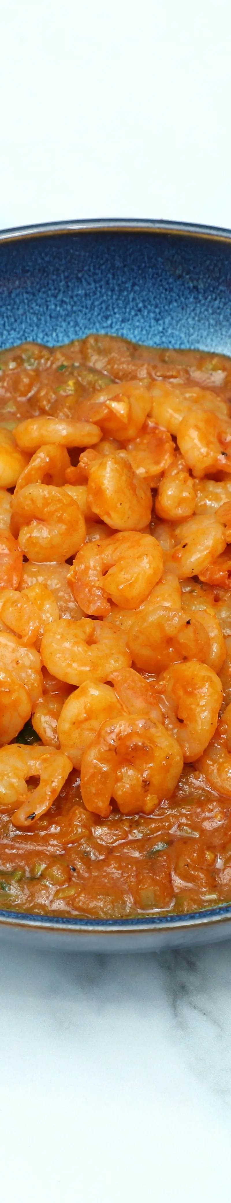 A blue bowl holds cooked king prawns in a vibrant orange Balti sauce on a marble surface.