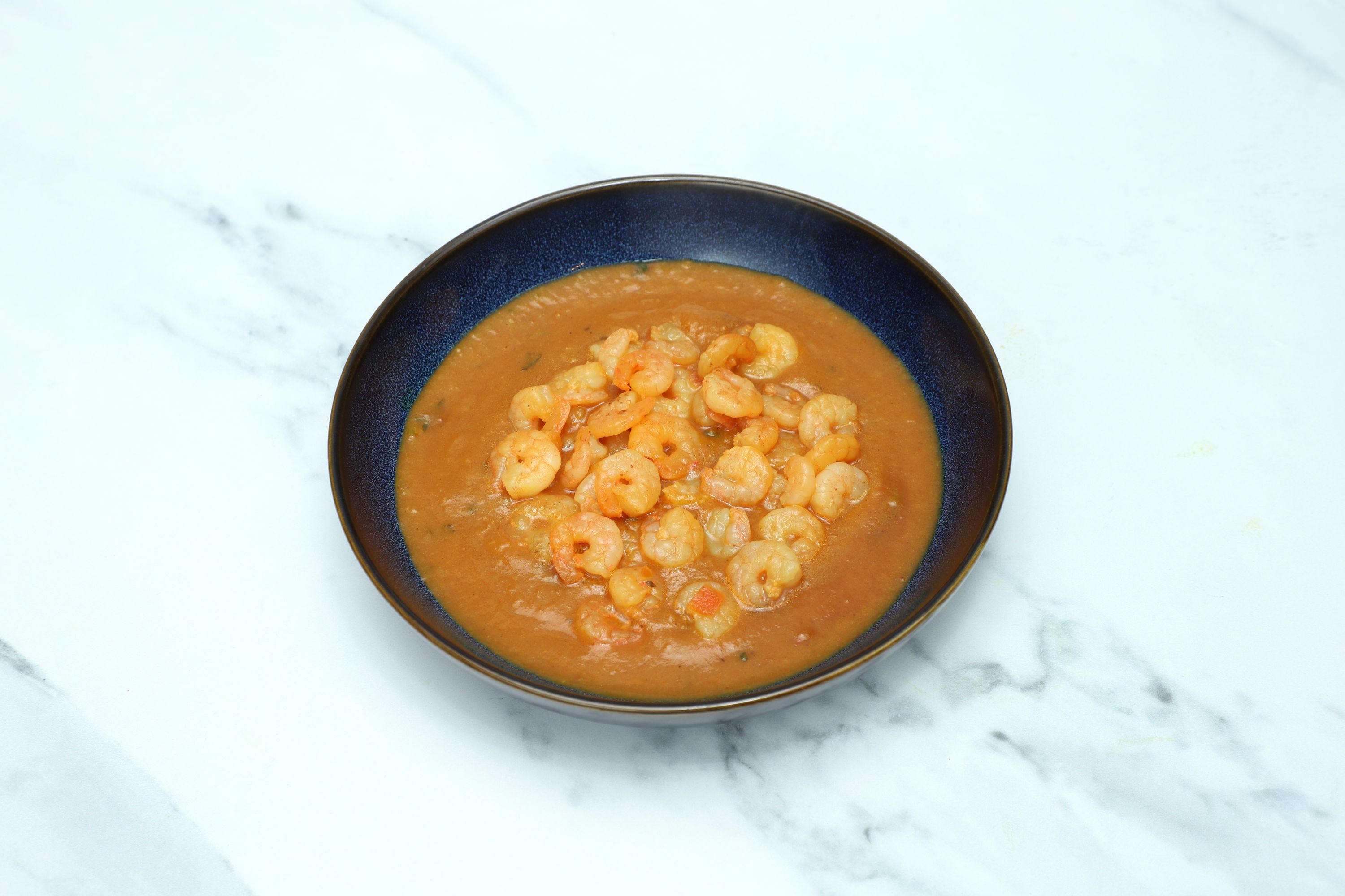 Shrimp curry with an orange-brown sauce and many small shrimp in a dark blue bowl.