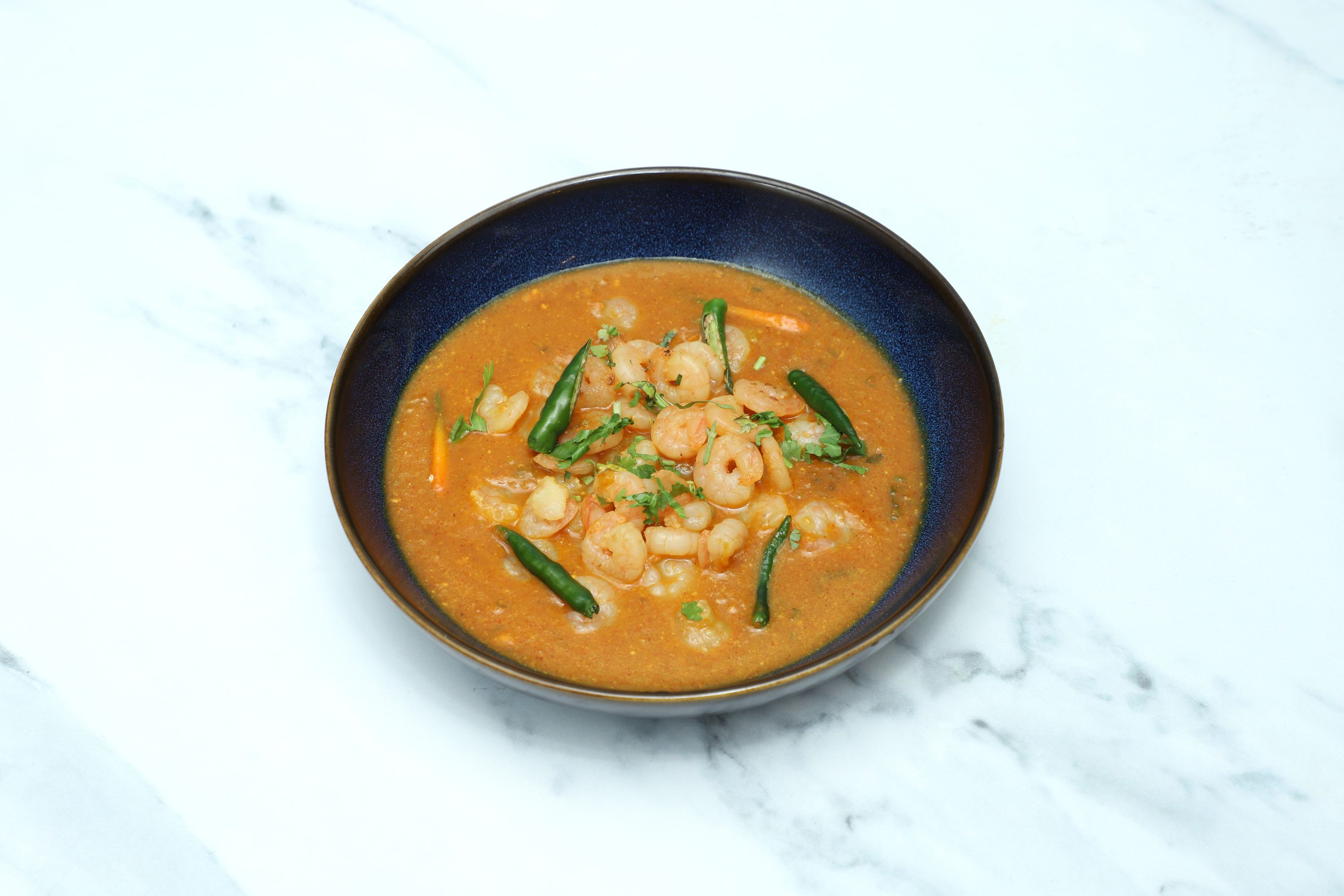 A dark blue bowl filled with orange prawn curry, garnished with green chillies and coriander.