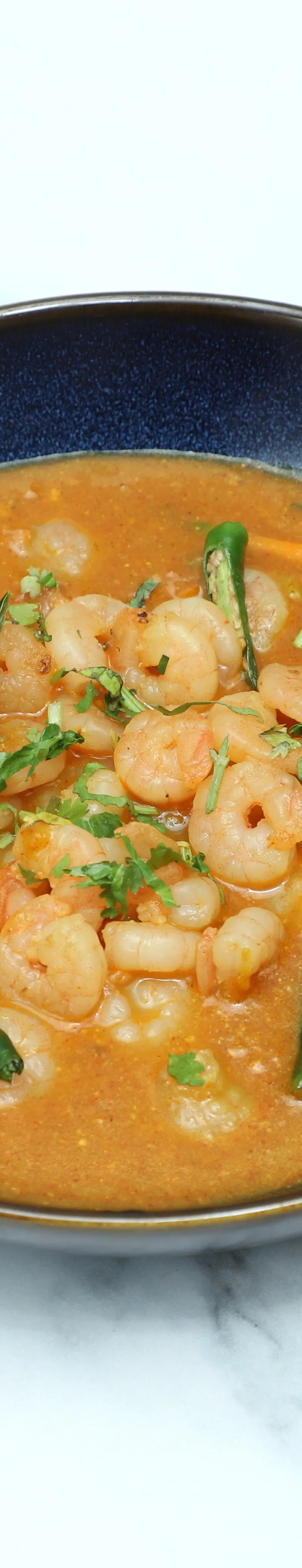 A dark blue bowl filled with orange prawn curry, garnished with green chillies and coriander.