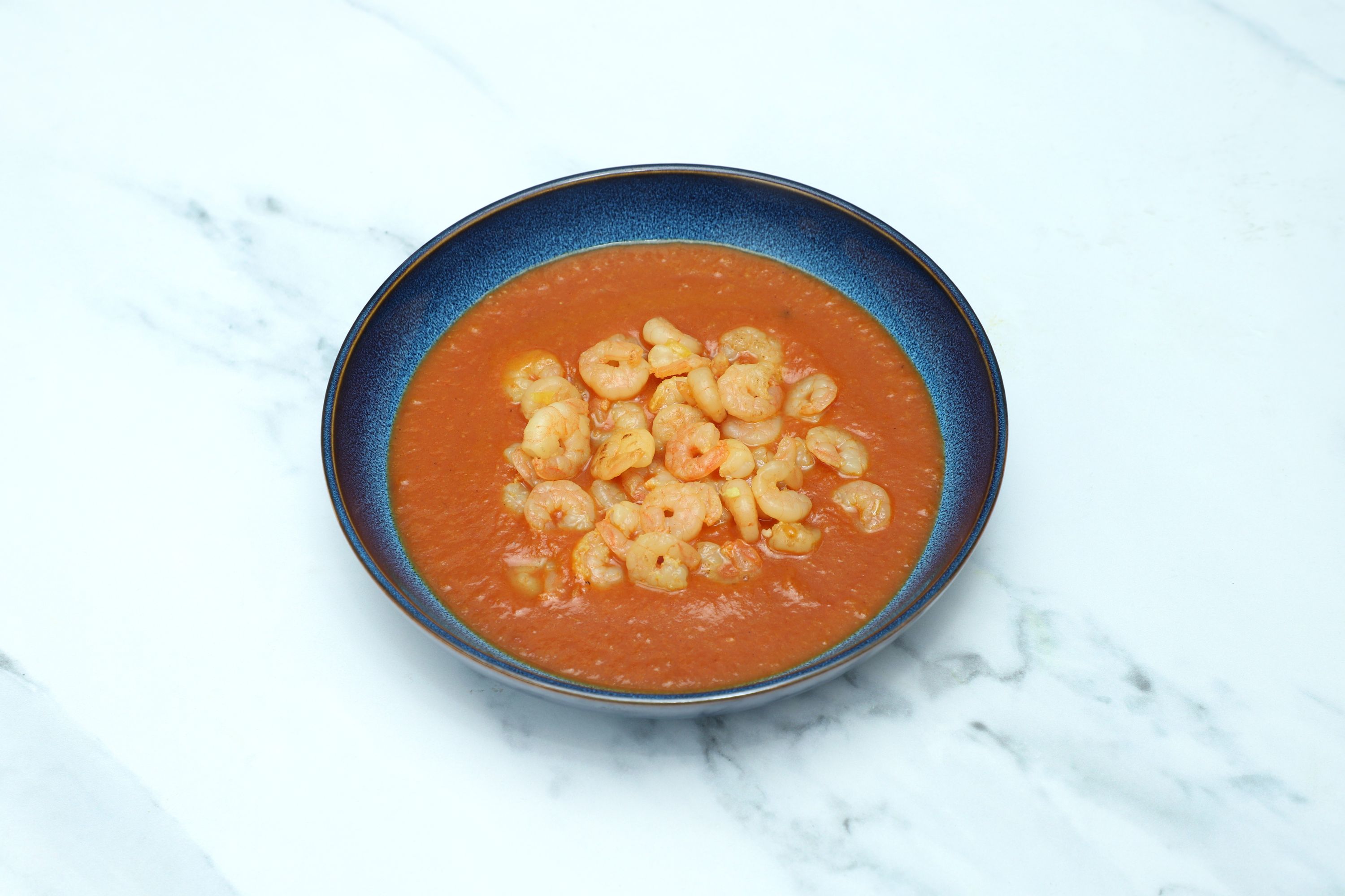 Top-down view of a dark blue bowl filled with orange-red curry and many small cooked shrimp.